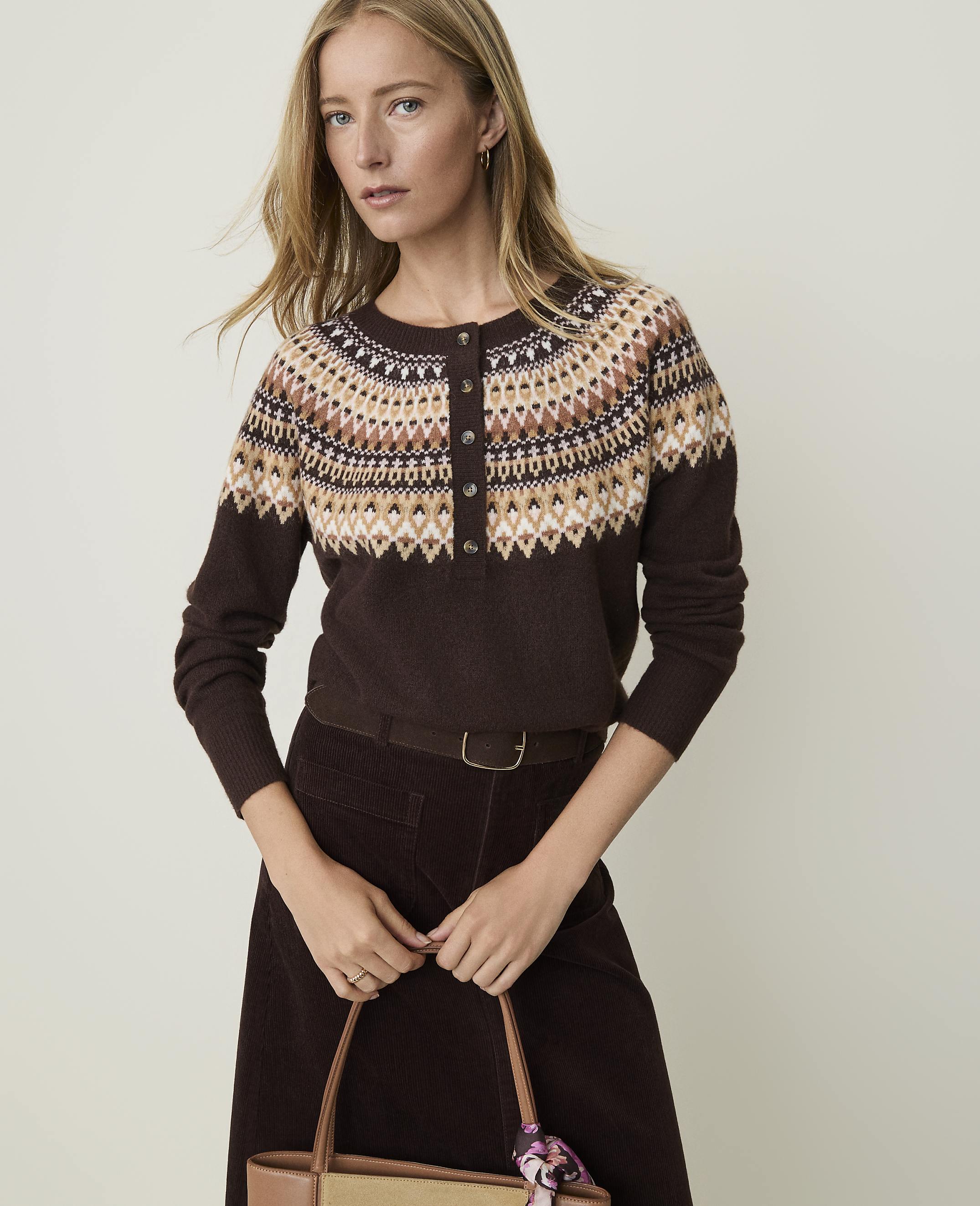 A woman in a brown fair isle sweater and corduroy skirt holds a tote bag in front of a plain, light-colored background.