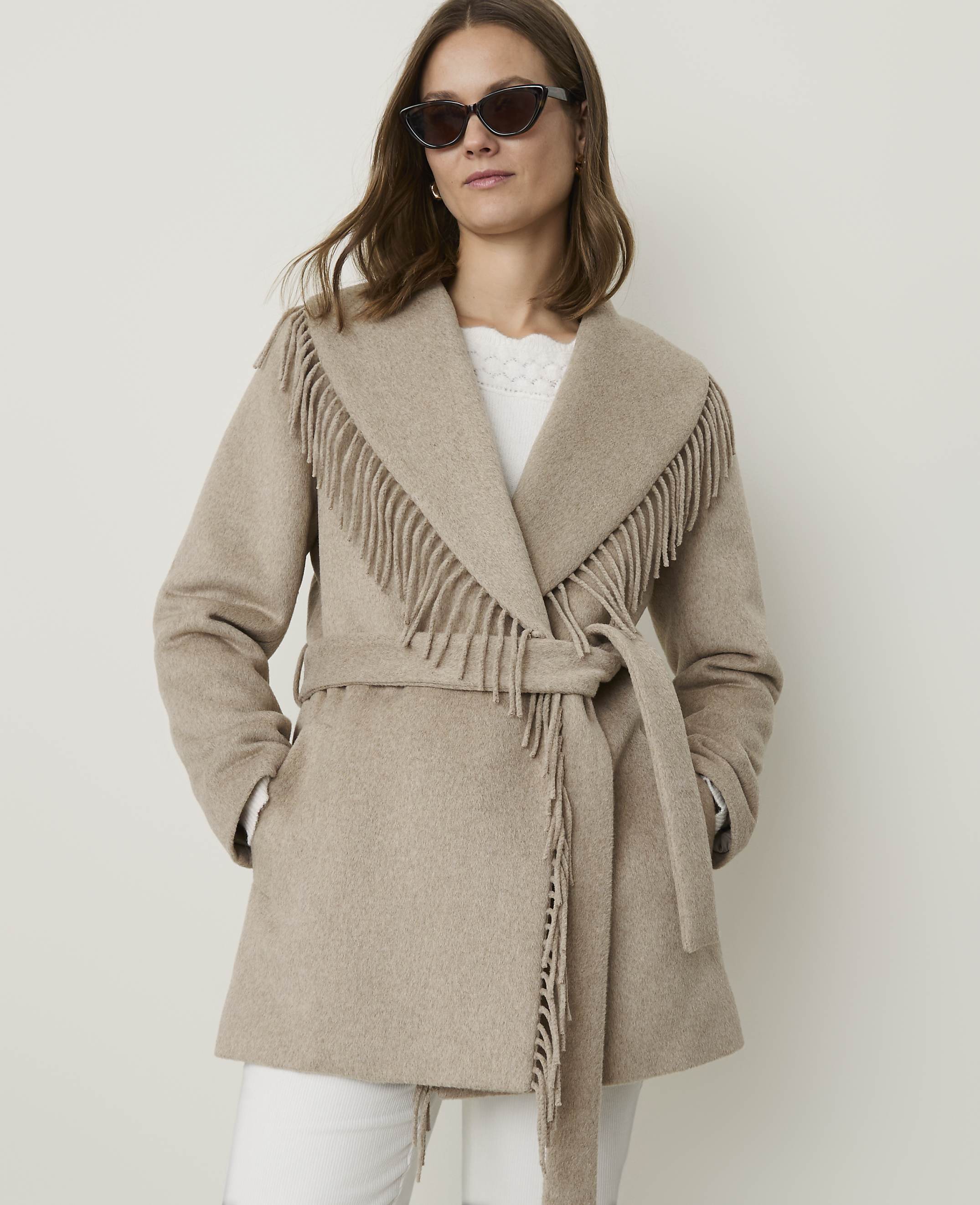 A woman in a fringed beige coat stands against a plain white background.