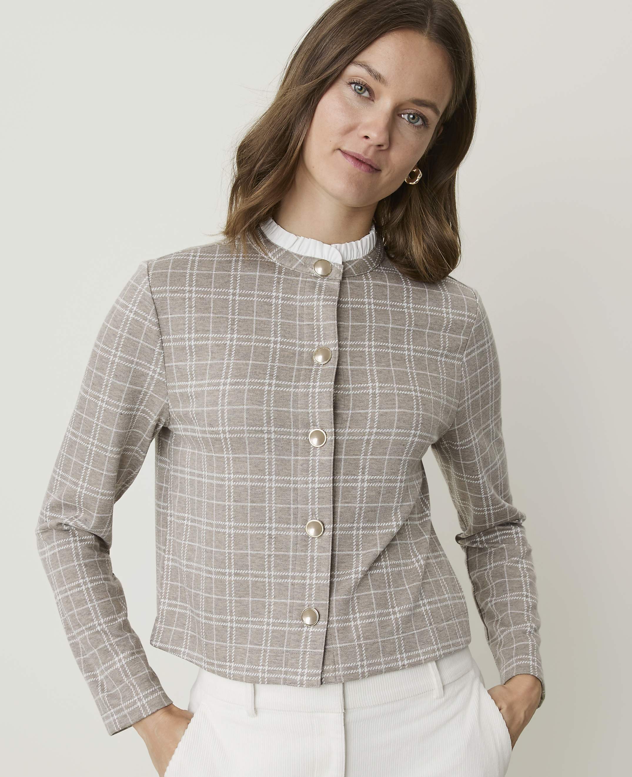 A woman in a plaid jacket and white pants stands against a plain background.