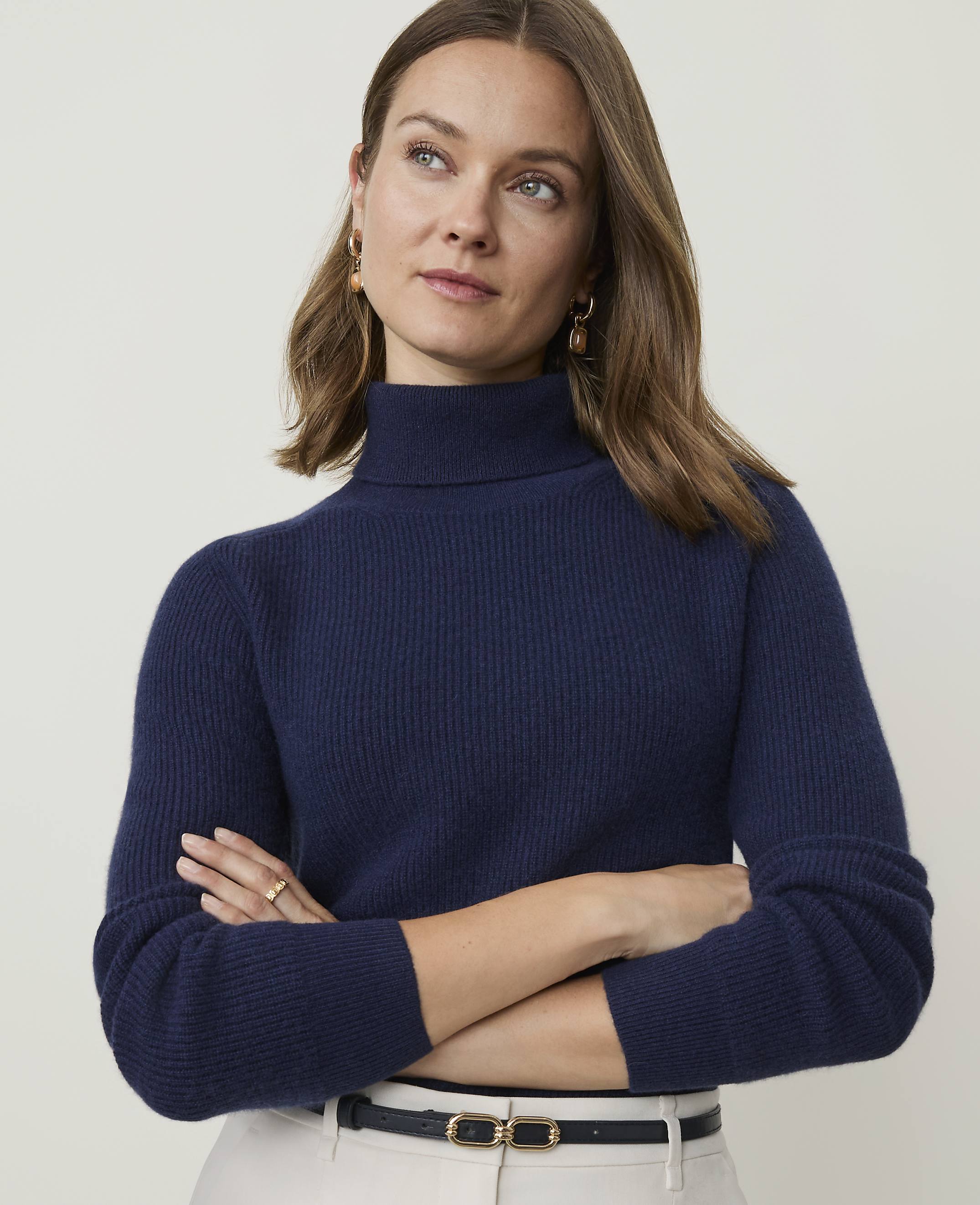 A woman in a navy turtleneck sweater and light-colored pants stands against a plain, light background.