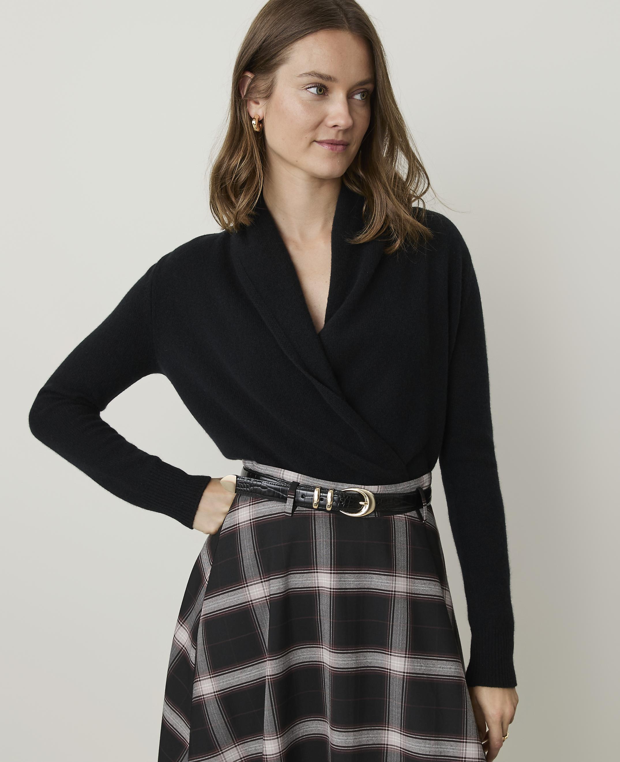 A woman in a black wrap sweater and plaid skirt stands against a plain, light background.