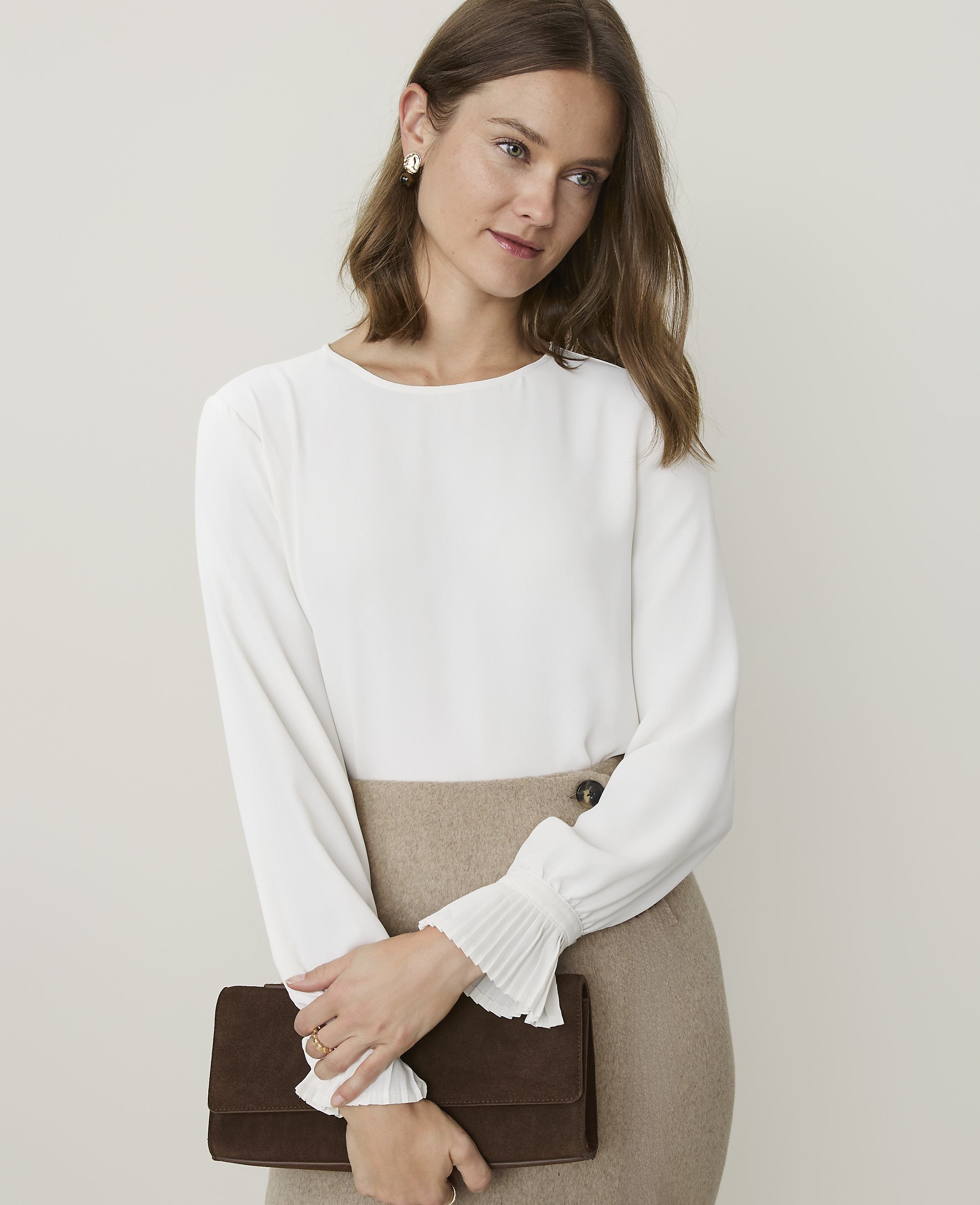 A woman in a white blouse and tan skirt holds a brown clutch in front of a plain background.
