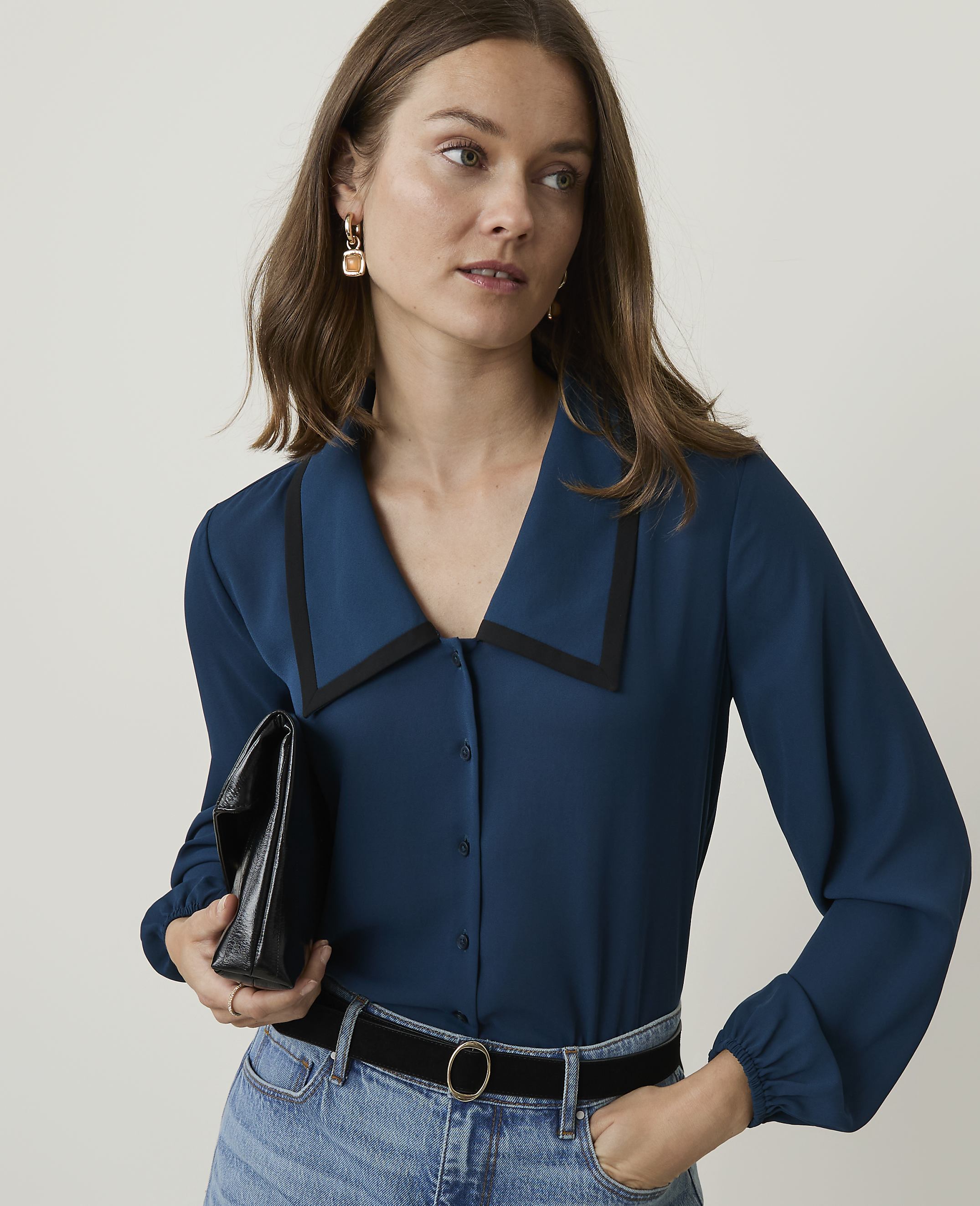 A woman in a teal blouse and jeans holds a black clutch in front of a plain background.