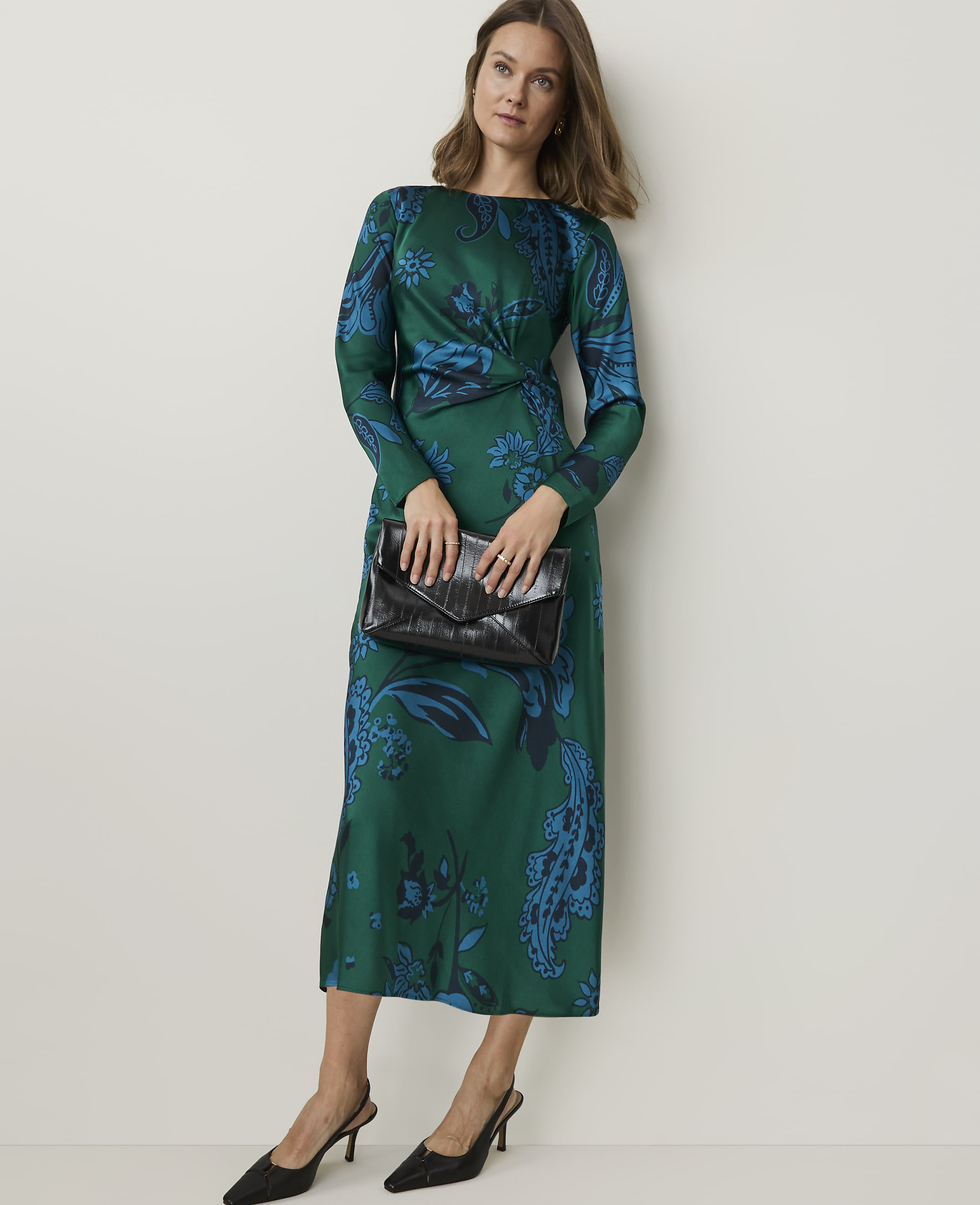 A woman in a green paisley dress and black heels holds a black clutch in front of a plain white background.