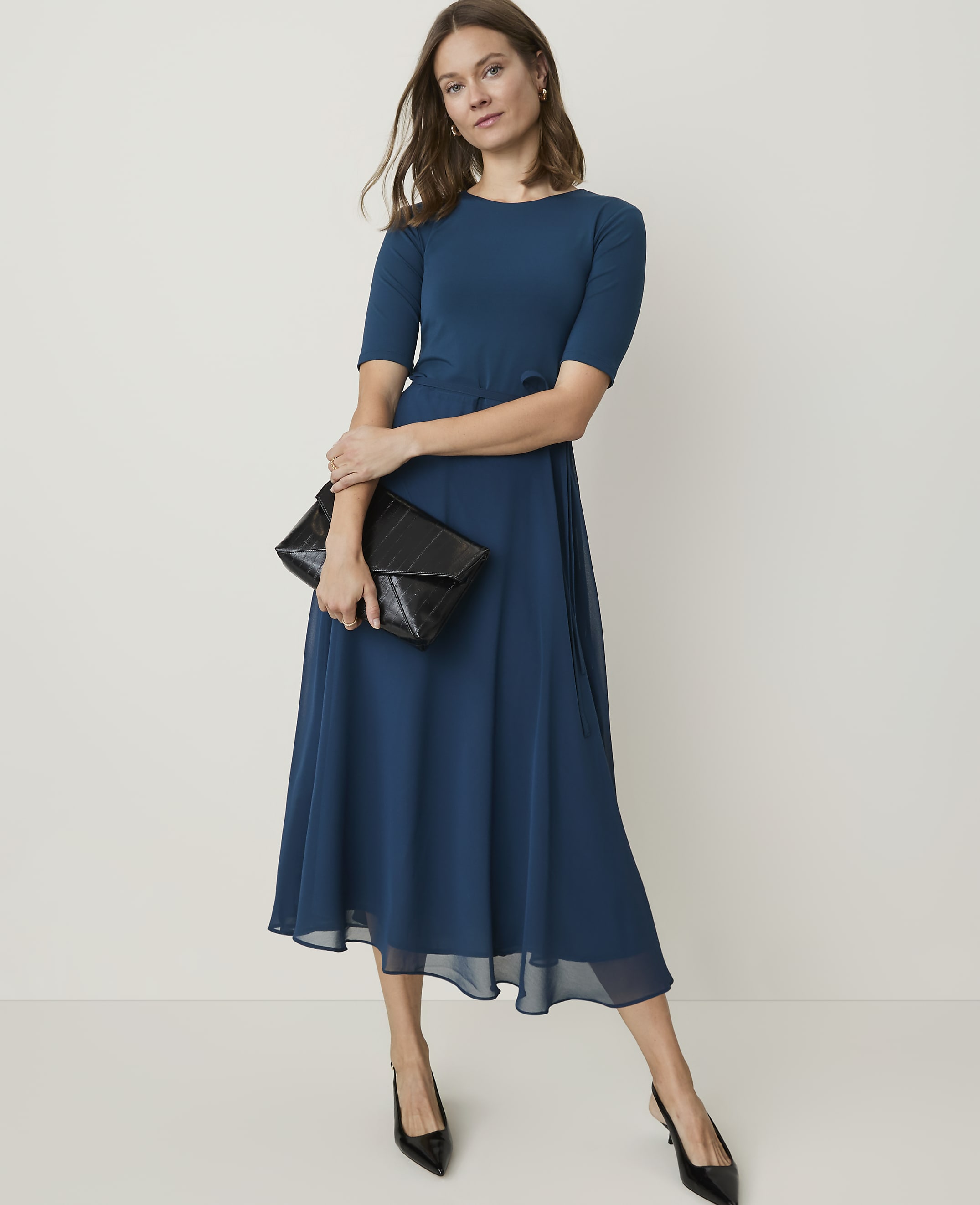 A woman in a teal dress and black heels holds a black clutch in front of a plain white background.