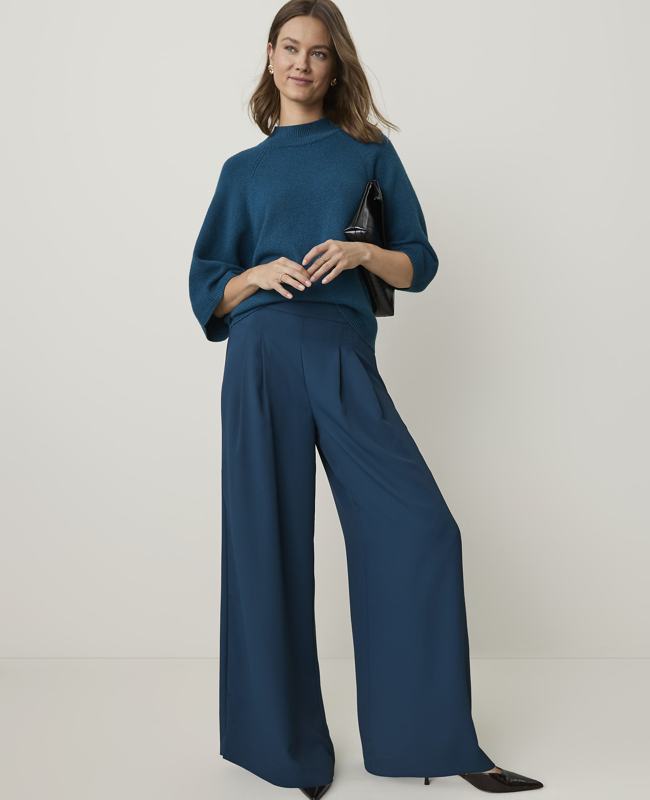 A woman in a teal sweater and wide-leg pants stands against a plain, light background, holding a black purse.
