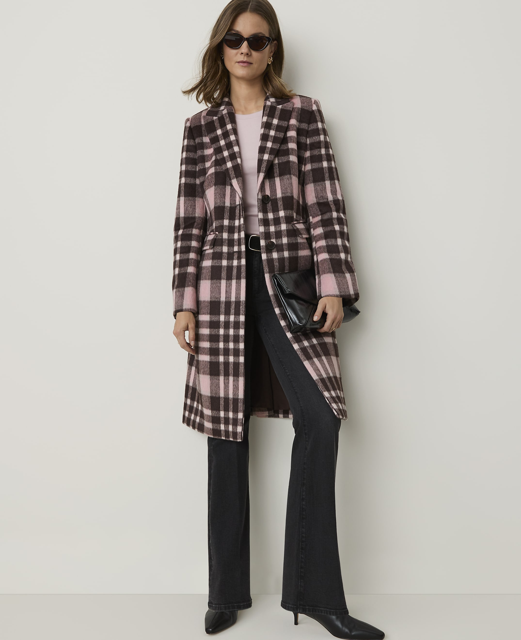 A woman in a pink and brown plaid coat and dark jeans stands against a plain, light-colored wall.
