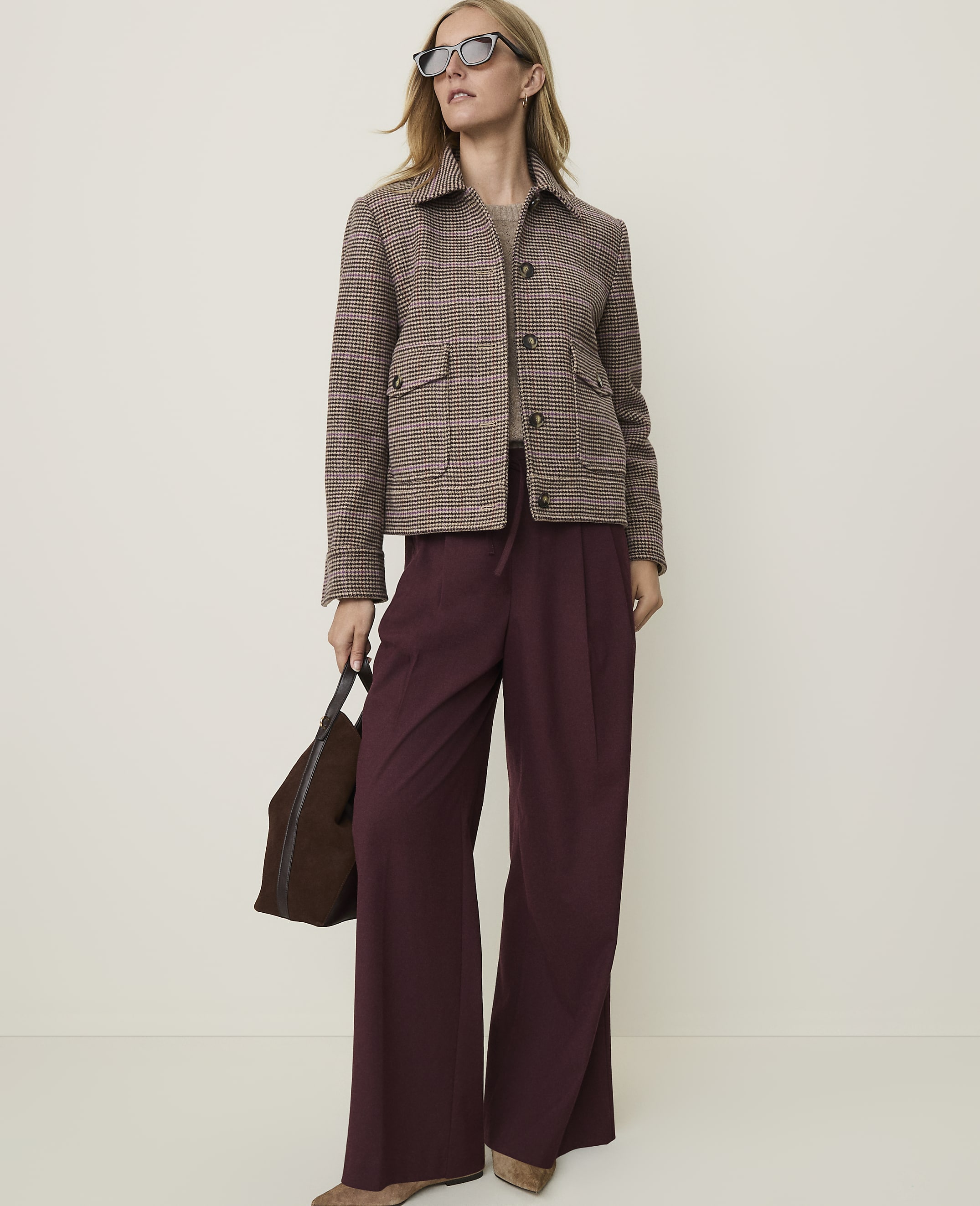 A woman in a brown plaid jacket and burgundy wide-leg pants stands against a plain, light-colored background, holding a dark brown handbag.