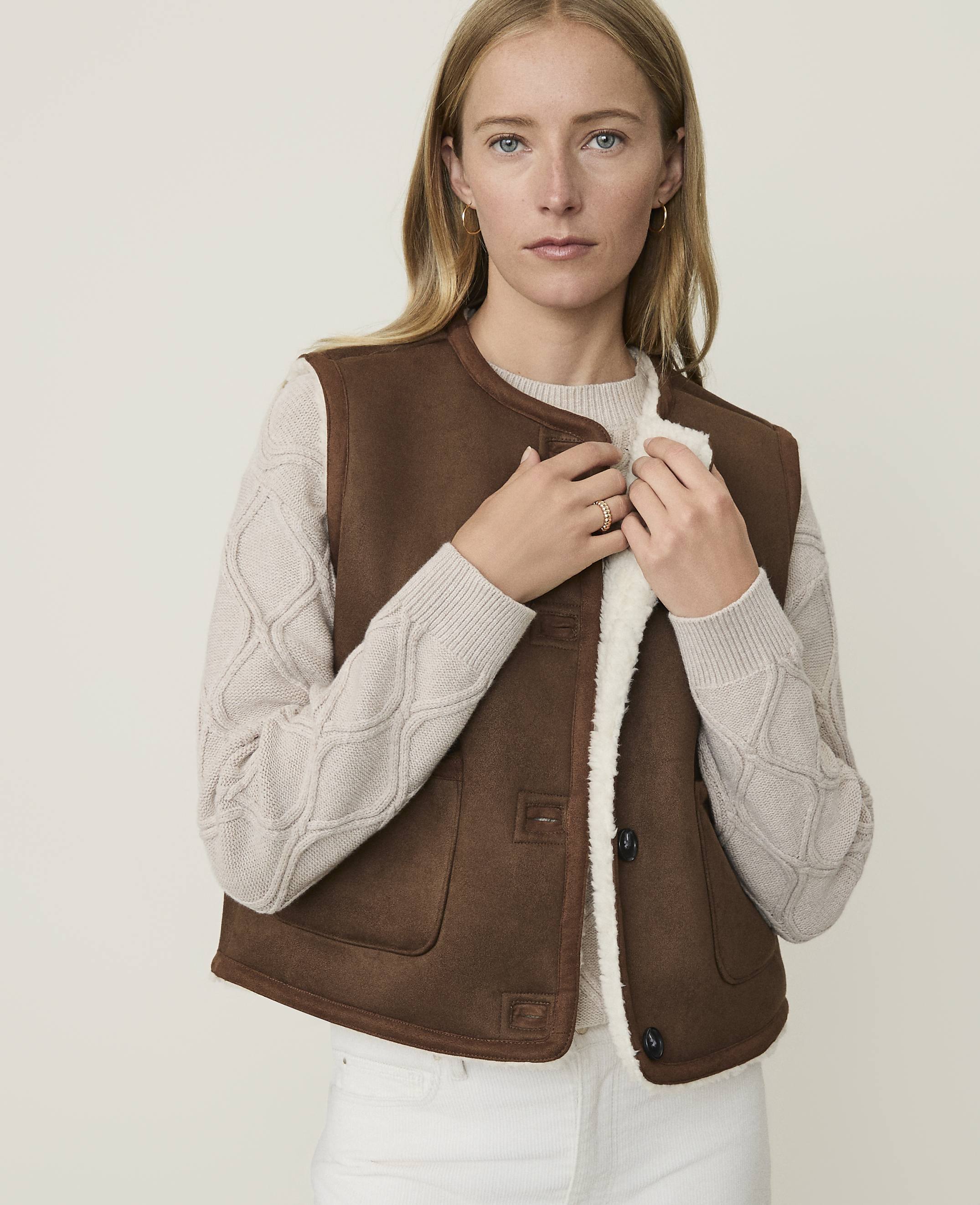 A woman in a brown shearling vest and cream sweater stands against a plain, light background.