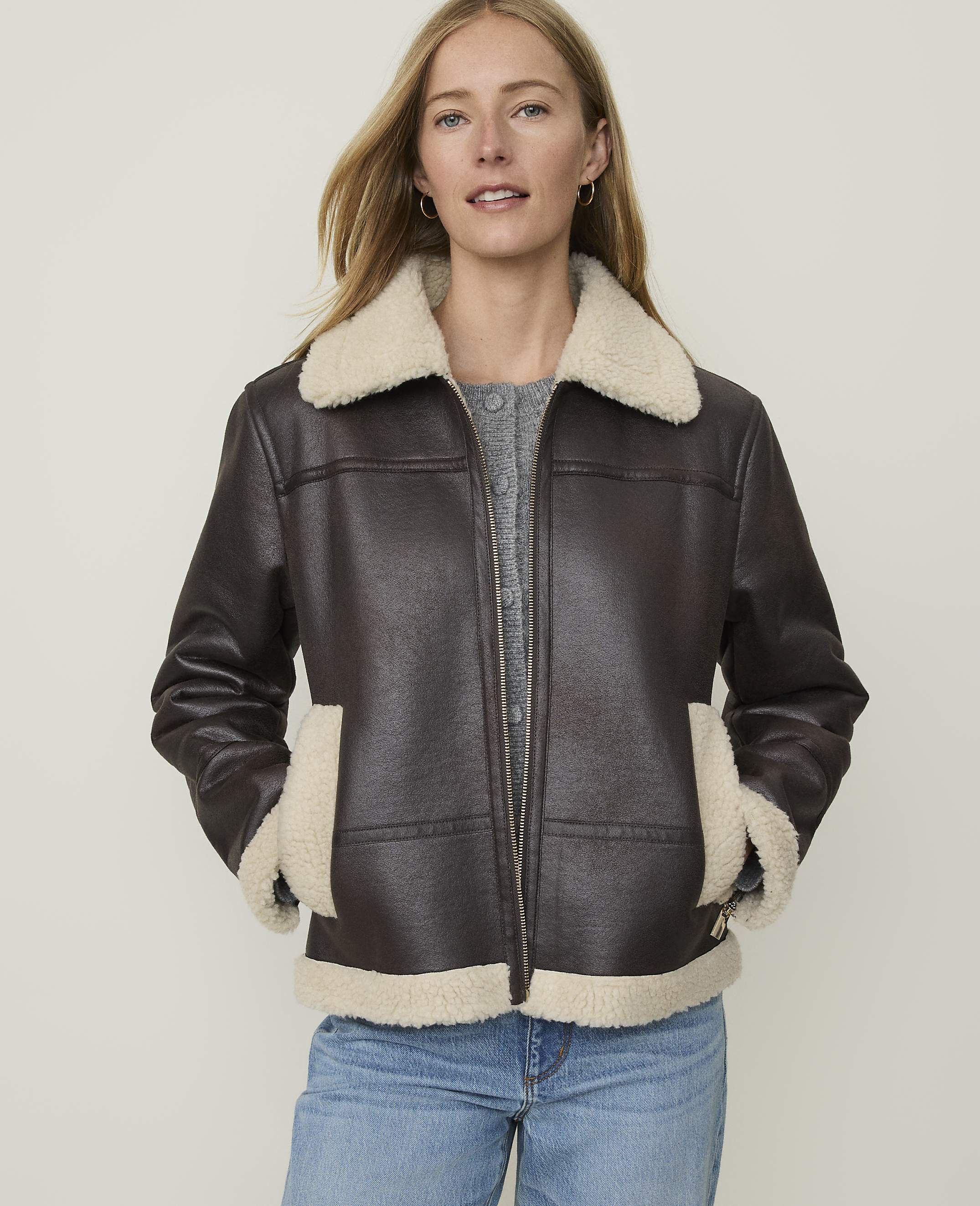 A woman in a brown faux-leather jacket with a cream shearling collar and cuffs stands against a plain, light background.