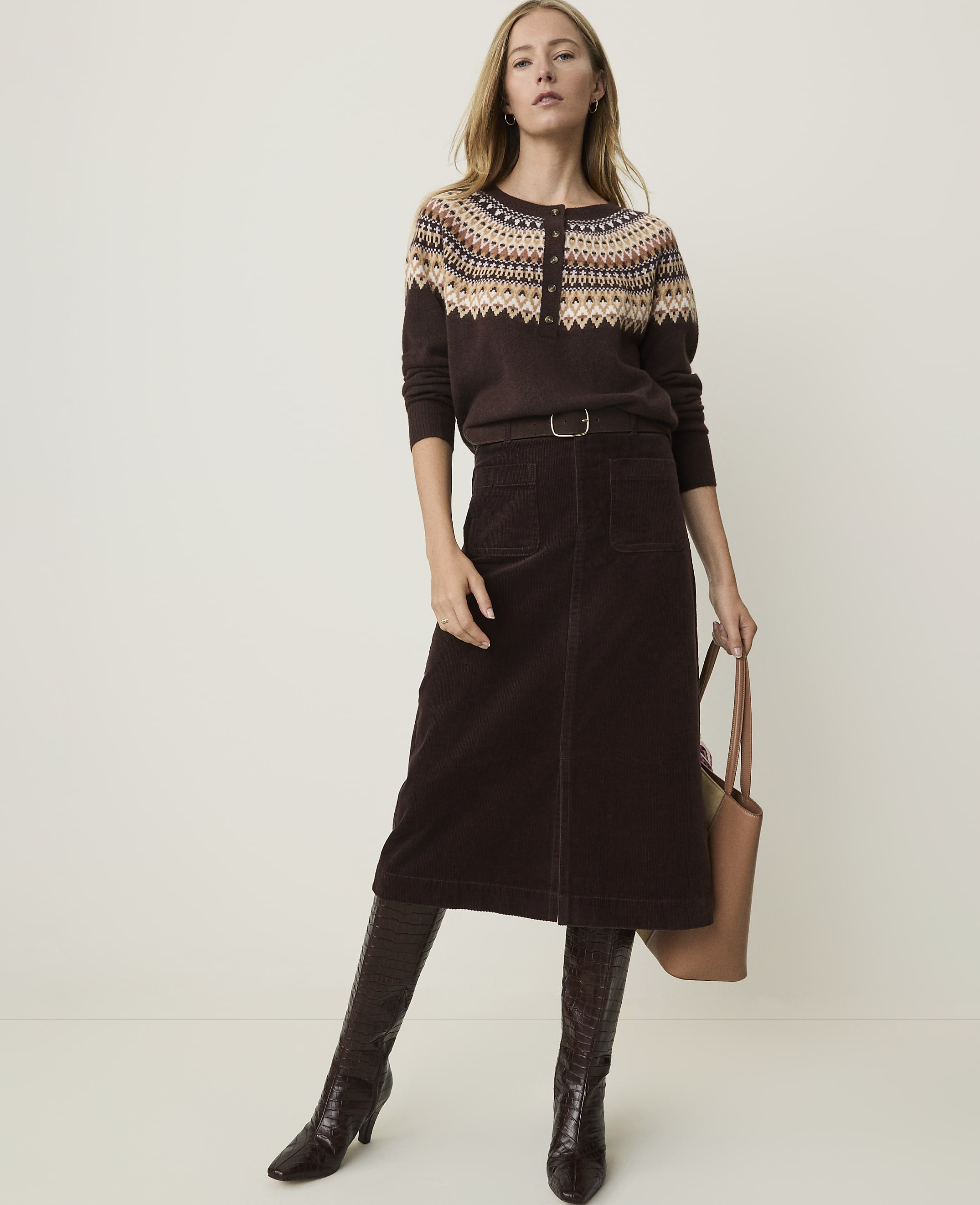 A woman in a patterned sweater, brown corduroy skirt, and croc-embossed boots stands against a plain, light-colored background, holding a tan tote bag.