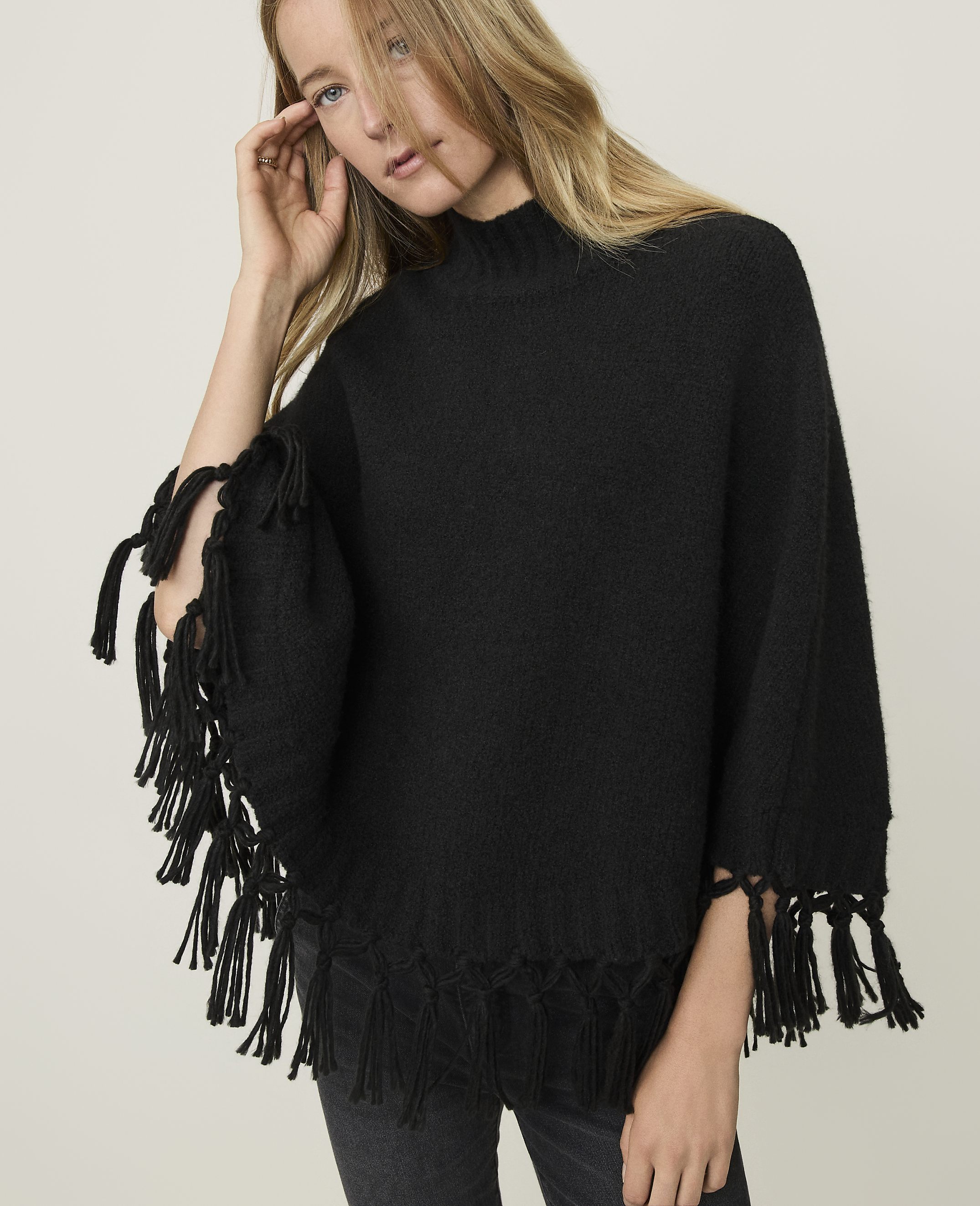 A woman in a black fringed poncho stands against a plain, light-colored background.
