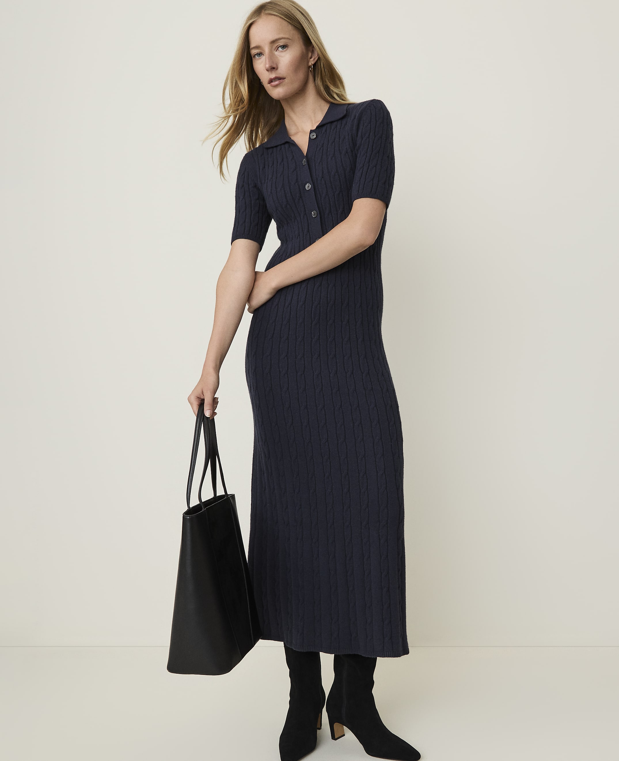 A woman in a navy cable-knit midi dress and black boots holds a black tote bag against a plain, light background.