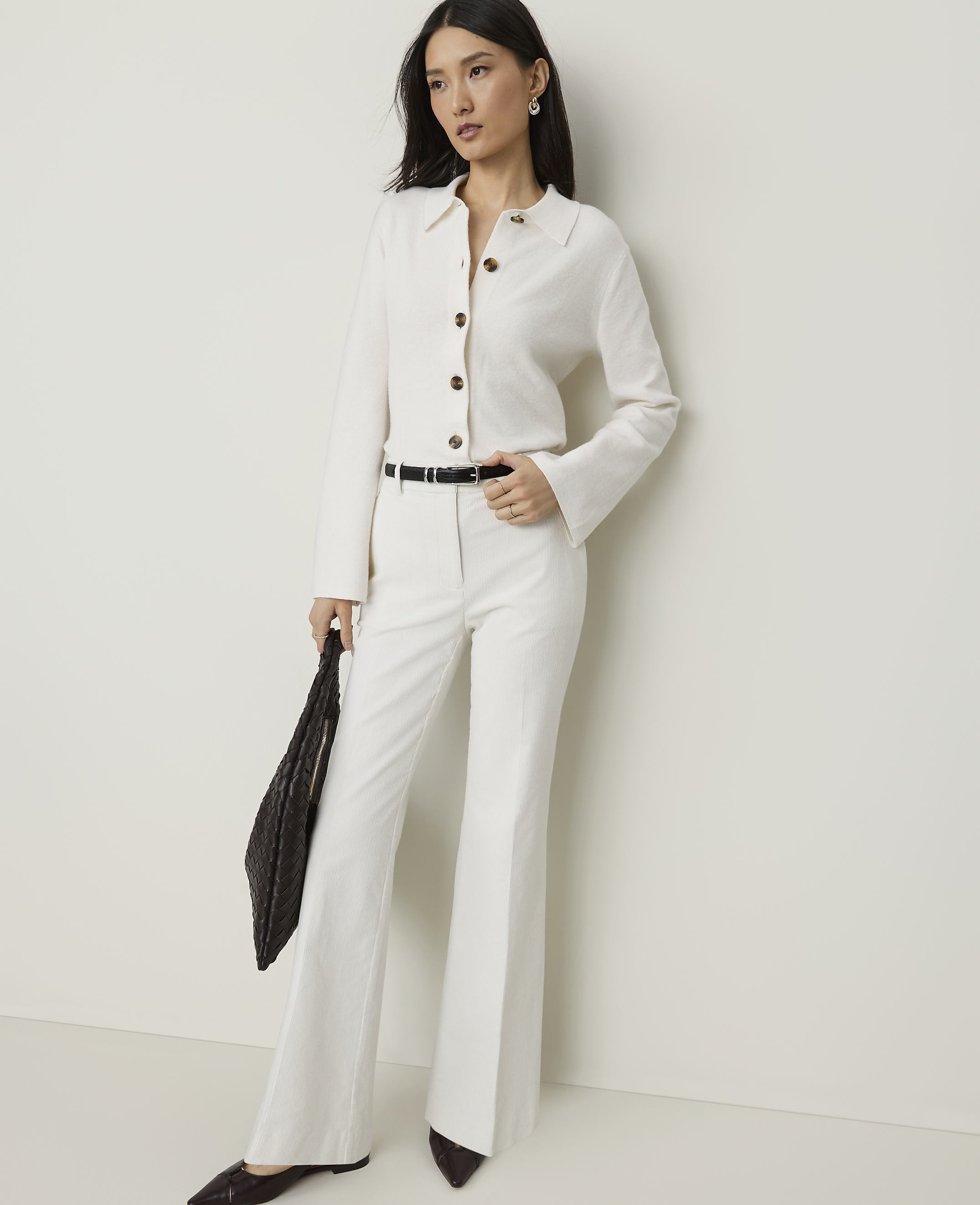 A woman in a white suit and dark shoes stands against a plain, light-colored wall, holding a dark woven bag.