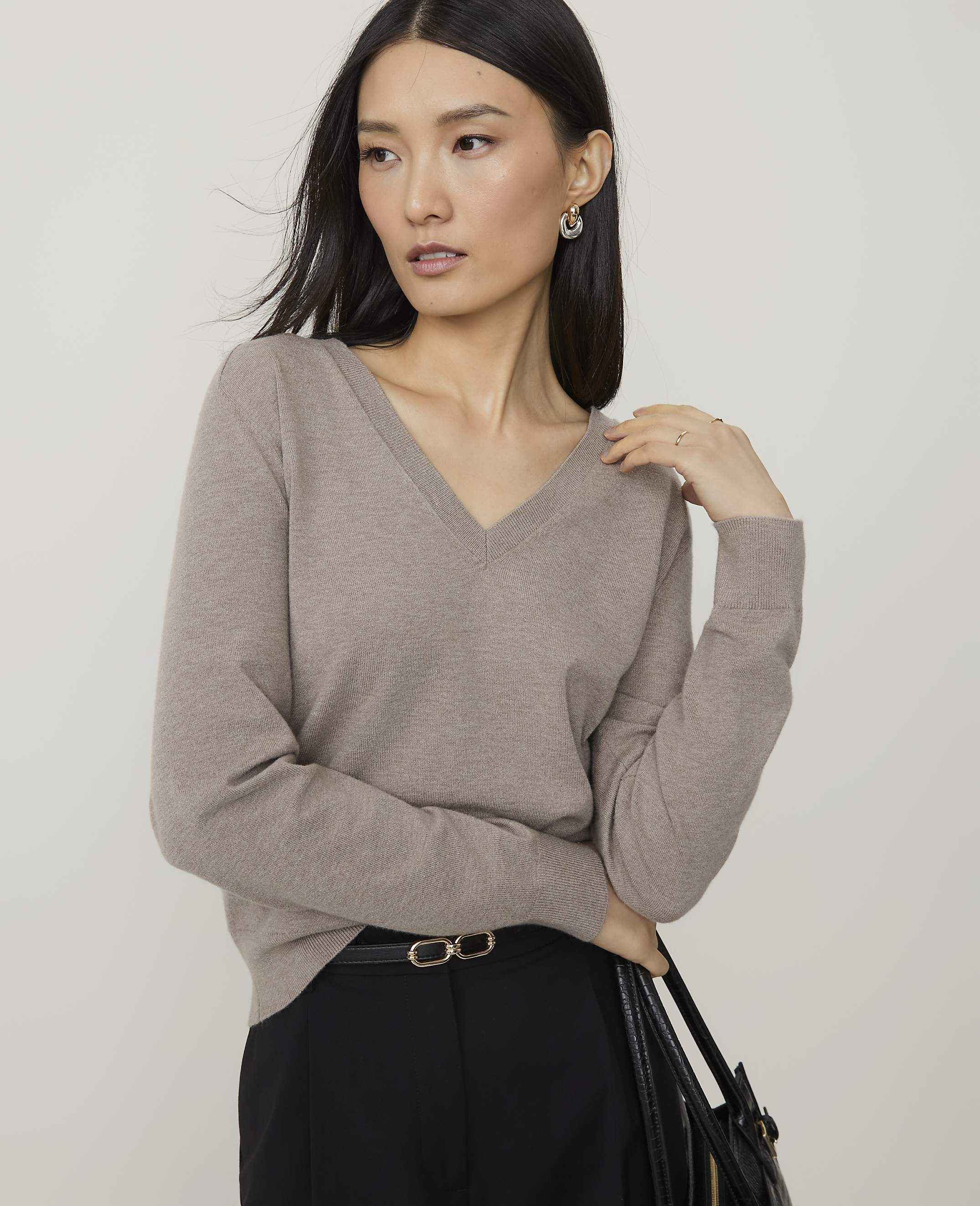 A woman in a taupe v-neck sweater and black pants stands against a plain gray background.