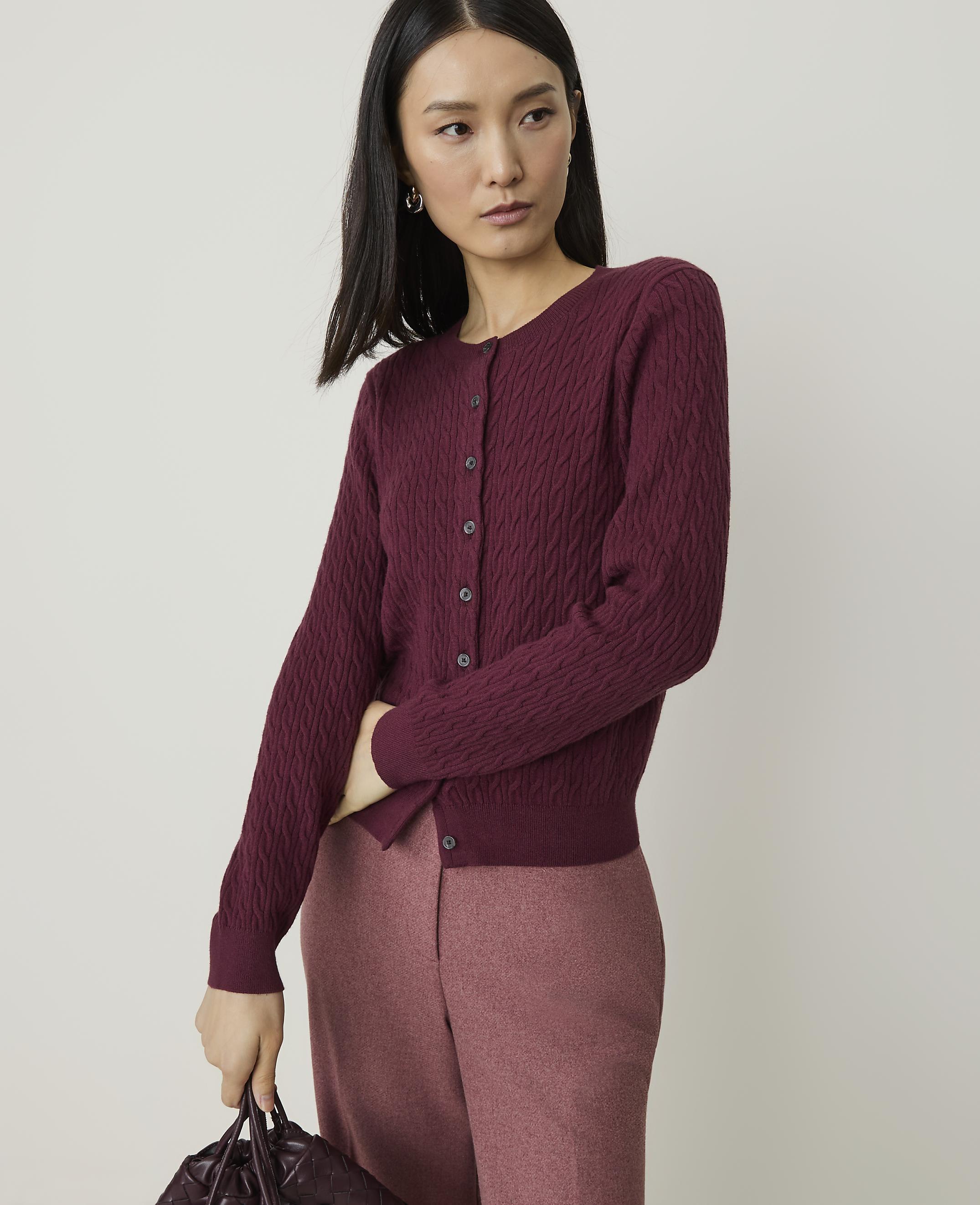 A woman in a burgundy cardigan and pink pants poses against a plain background, holding a dark handbag.