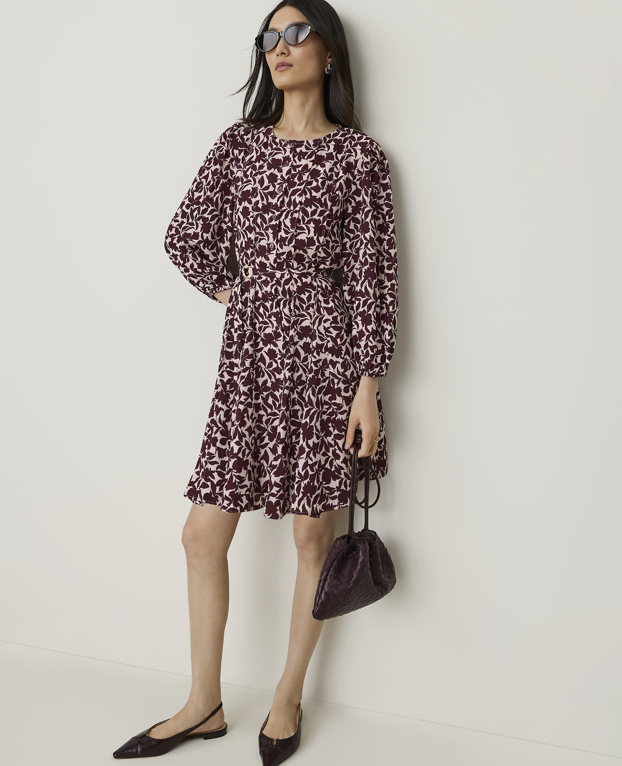 A woman in a floral dress and sunglasses stands with a handbag against a plain wall.
