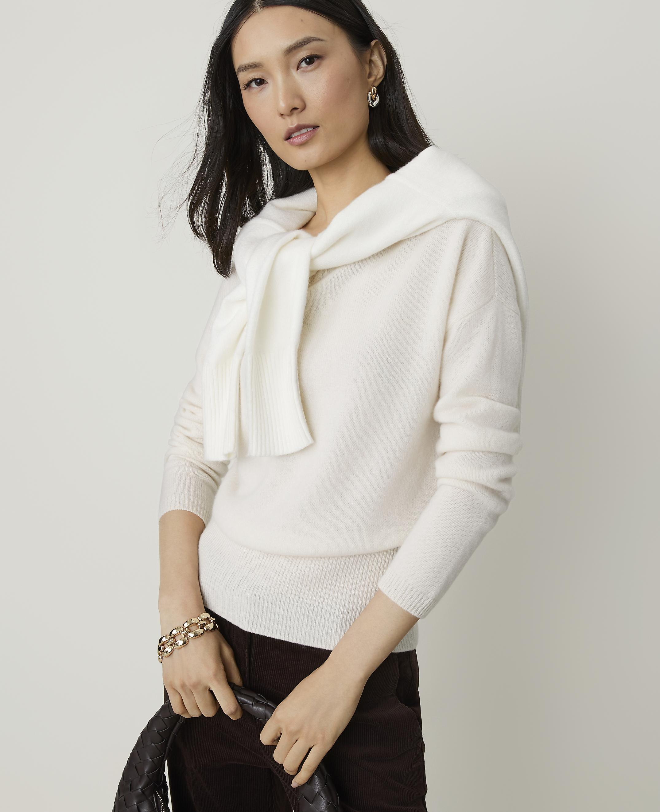 A woman in a cream sweater and dark pants holds a woven bag in front of a plain background.