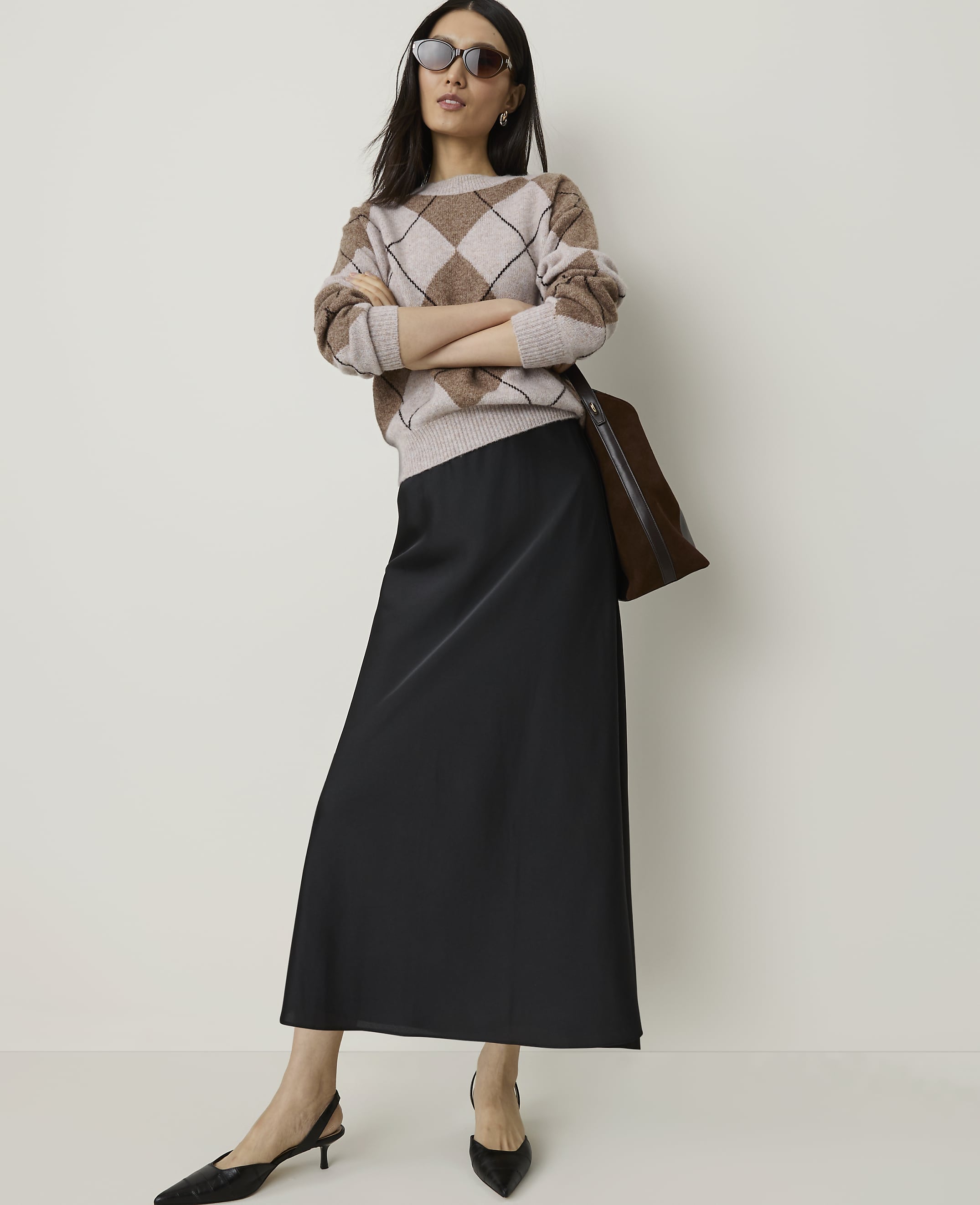 A woman in a beige argyle sweater and black slip skirt stands with her arms crossed against a plain, light-colored background.