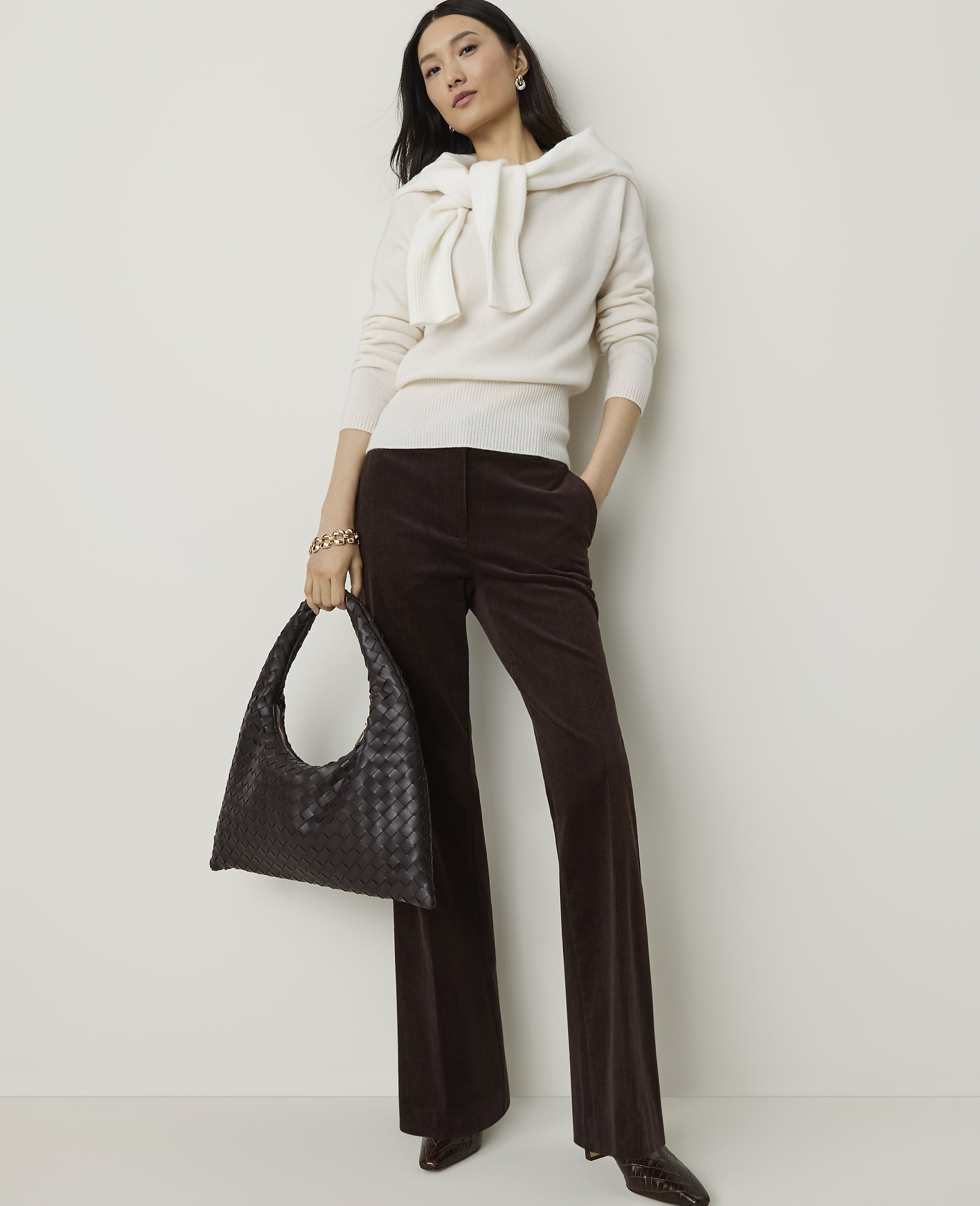 A woman in a cream sweater and brown corduroy pants stands with a dark woven bag against a plain white background.