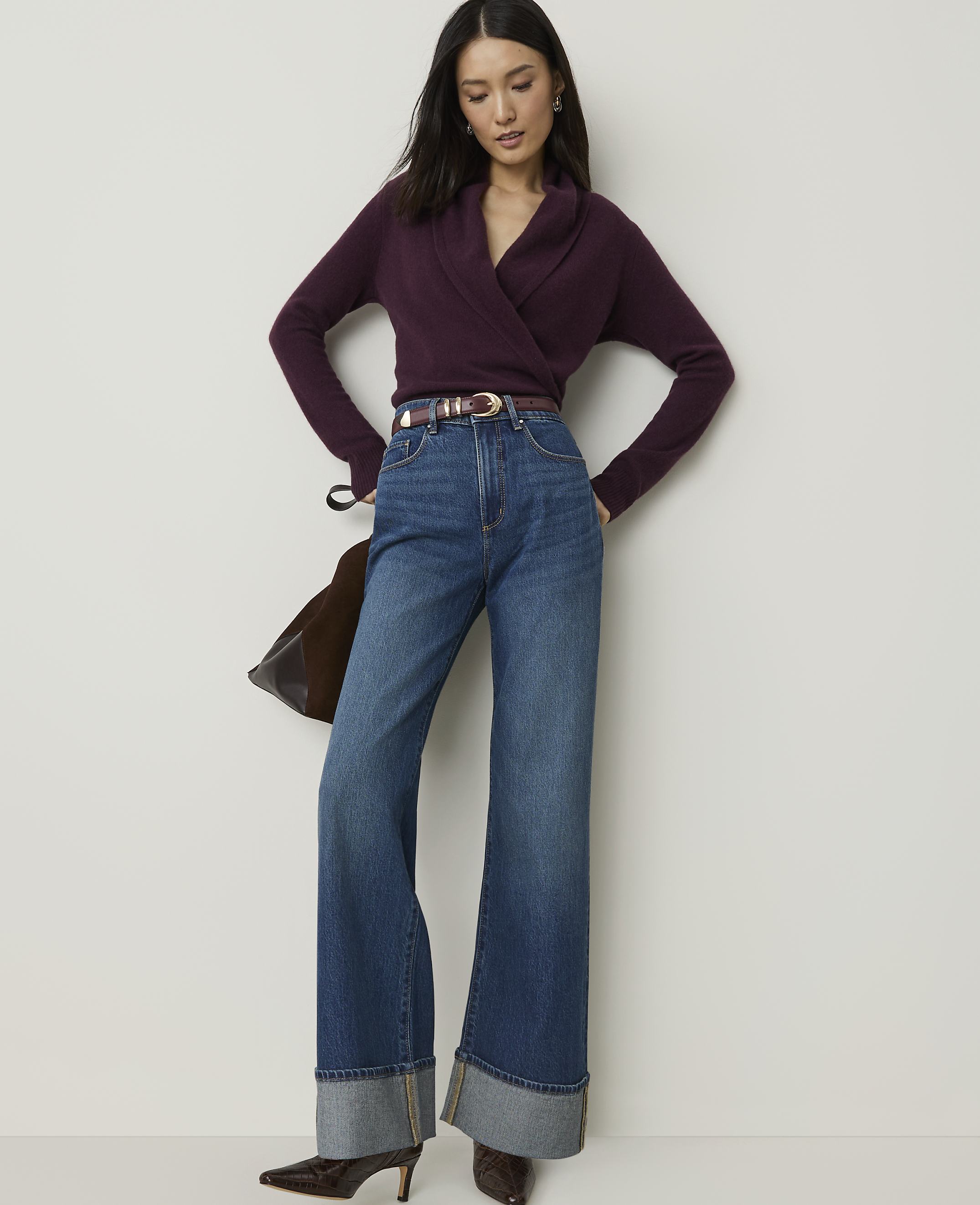 A woman in a plum-colored wrap sweater and wide-leg jeans stands against a plain white background.