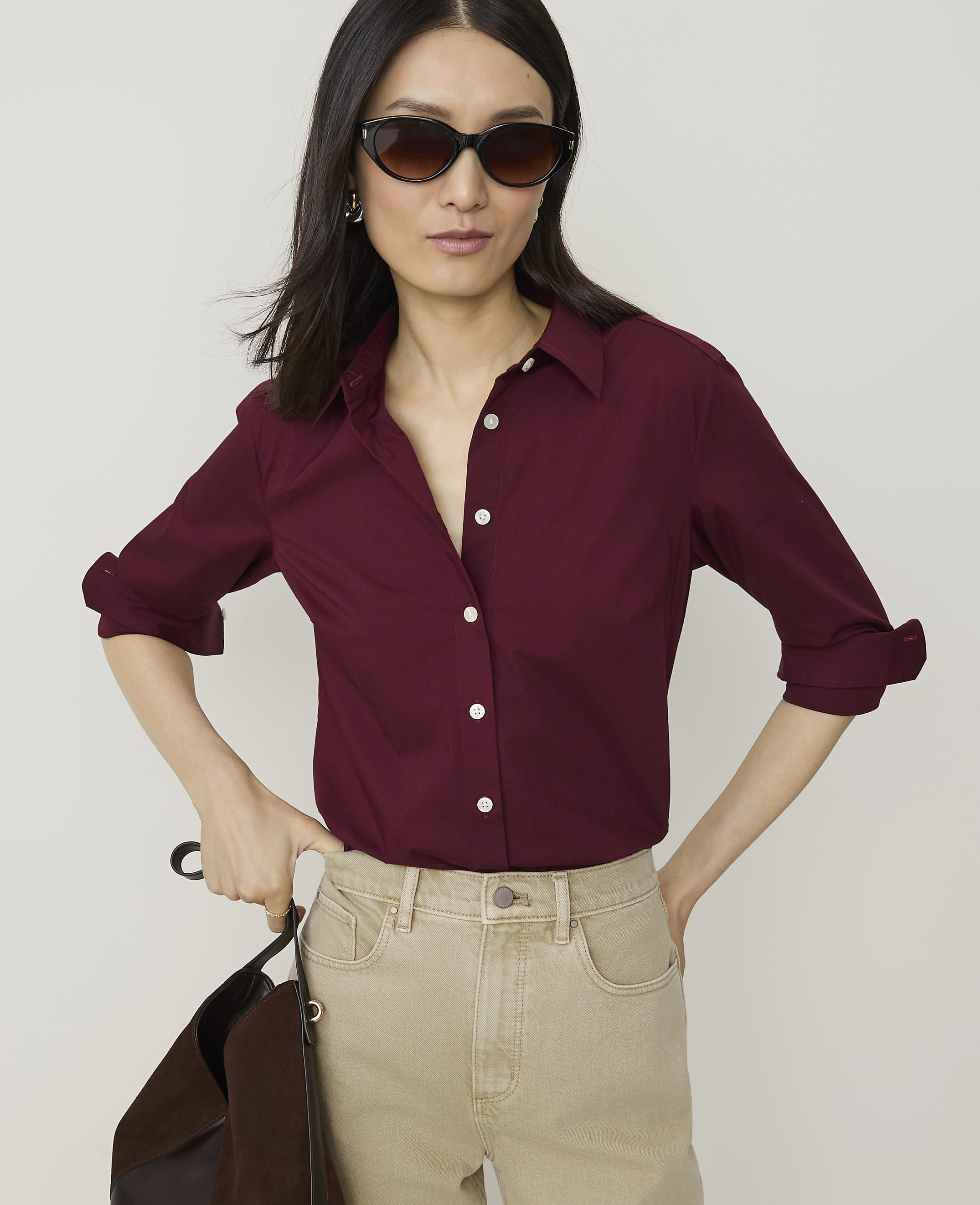A woman in a burgundy button-down shirt and tan pants stands against a plain white background, holding a dark brown bag.