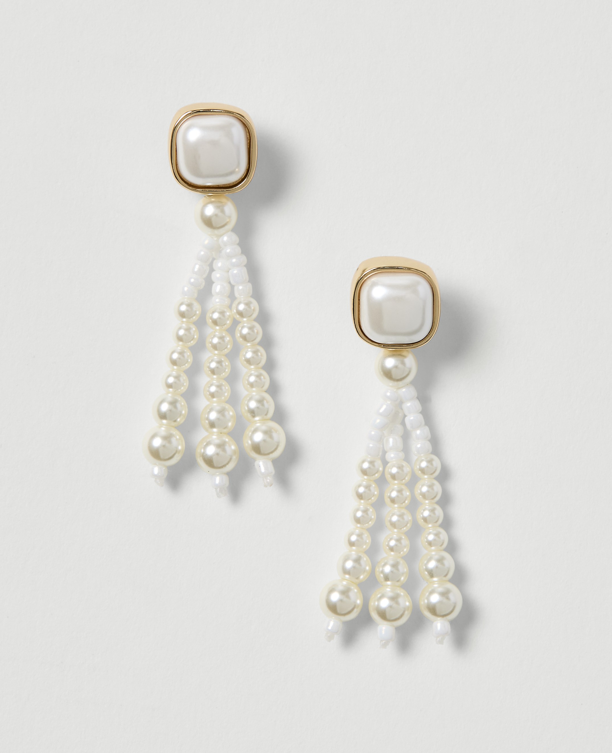 A pair of elegant pearl drop earrings with gold-toned square tops hang against a plain white background.
