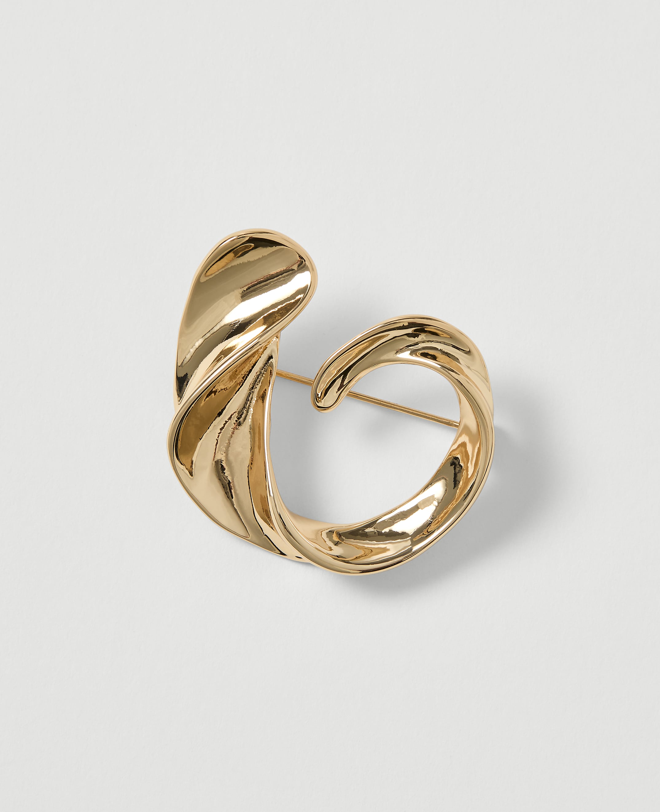 A polished gold abstract brooch with a pin is centered on a plain white background.