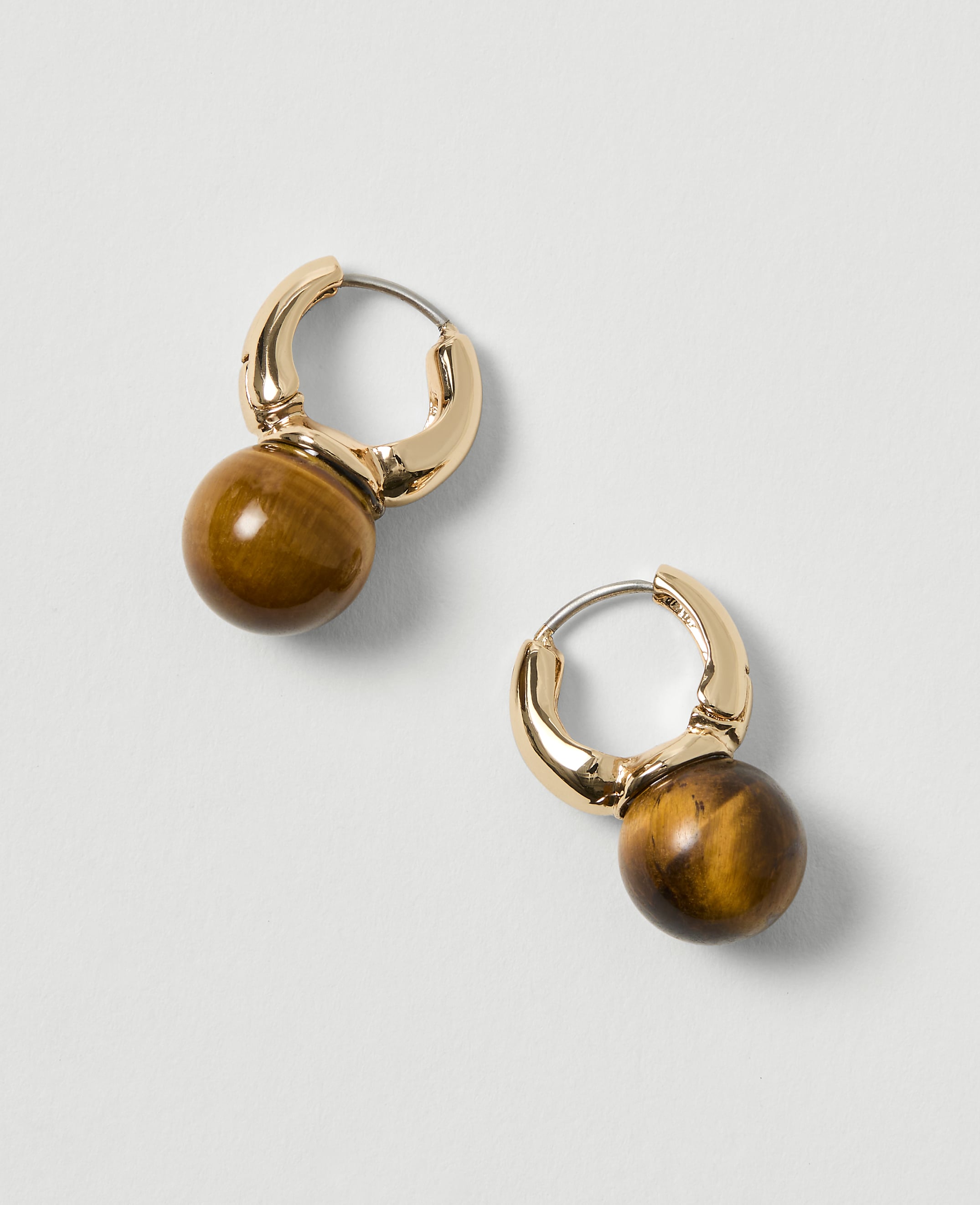 A pair of gold hoop earrings with tiger's eye beads hang against a plain, light gray background.