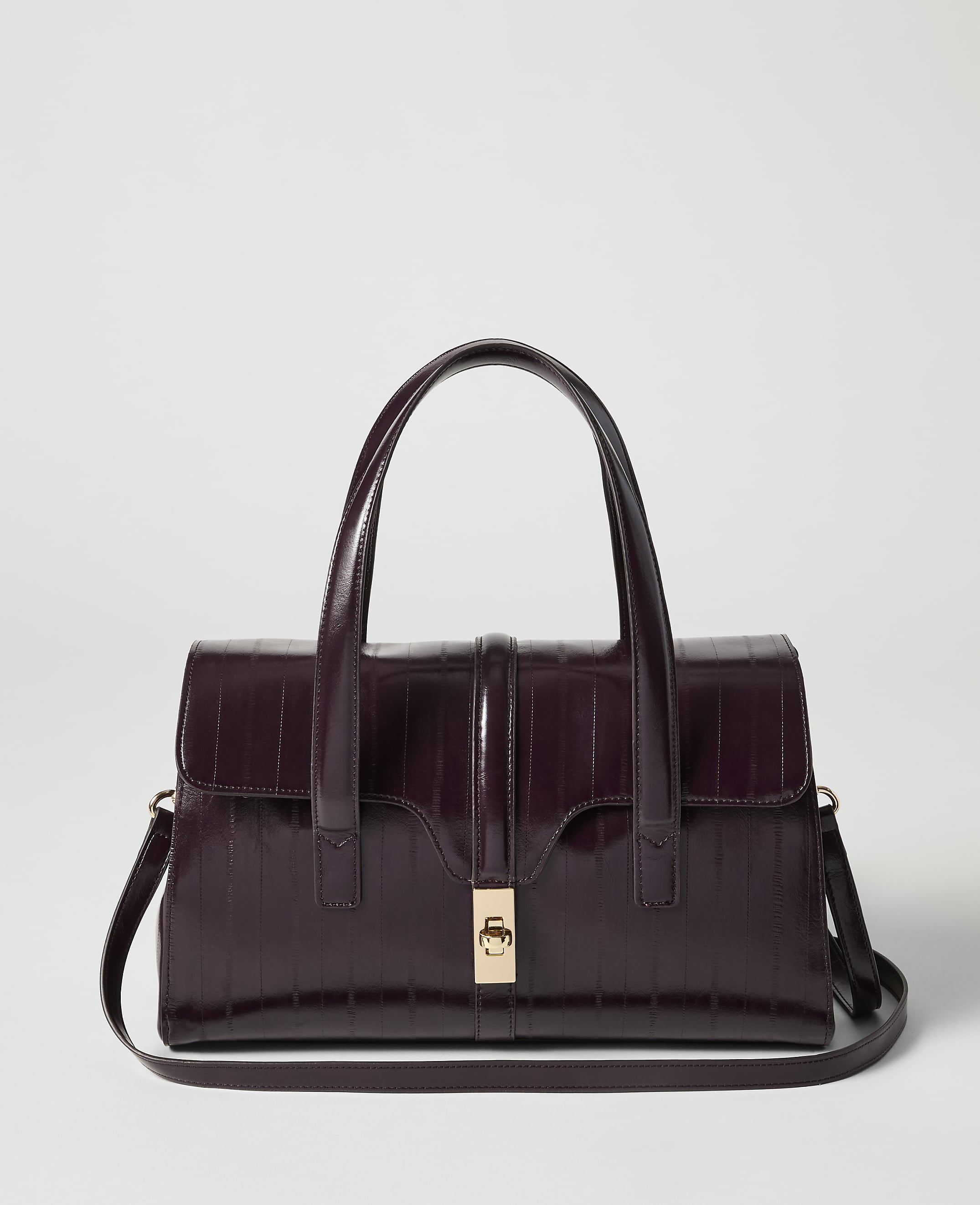 A deep purple, croc-embossed handbag with a gold clasp and top handles sits in the foreground against a plain, light gray background.