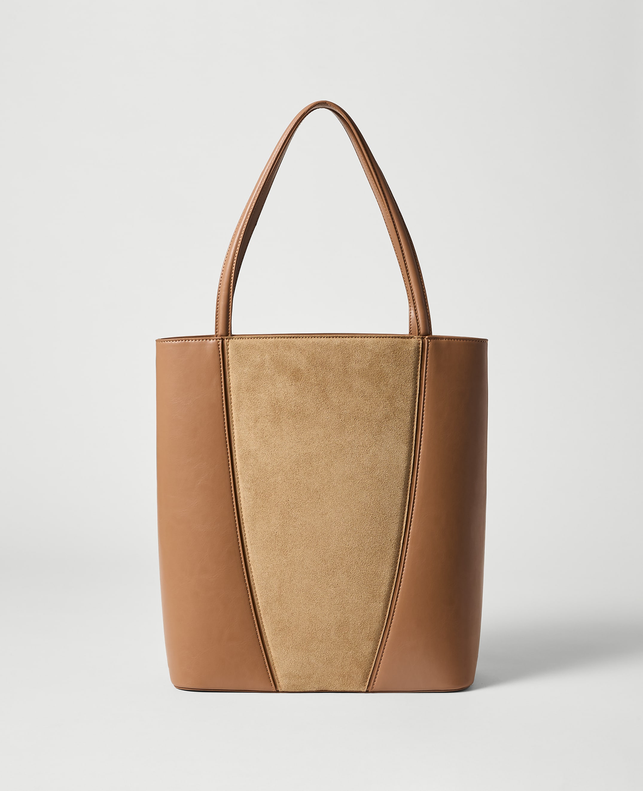 A tan leather and suede tote bag with thin straps stands against a plain, light gray background.