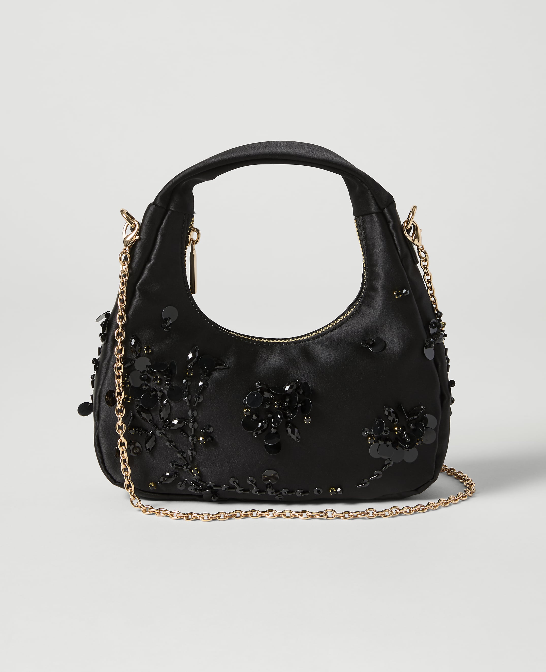 A black satin handbag adorned with black sequins and beads sits on a light gray background, with a delicate gold chain strap draped across it.