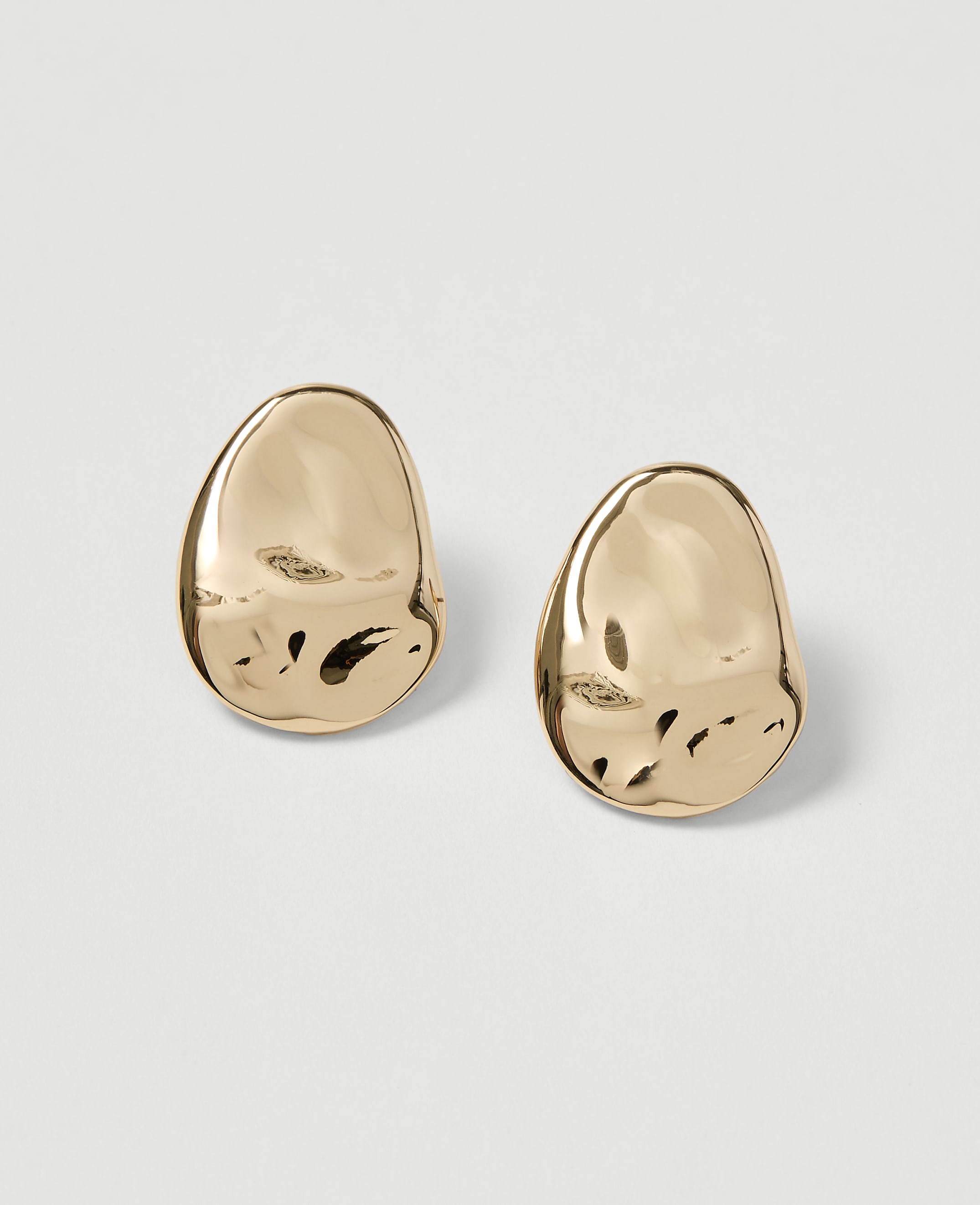 Two polished gold earrings with an organic, pebble-like shape rest on a plain, light gray background.
