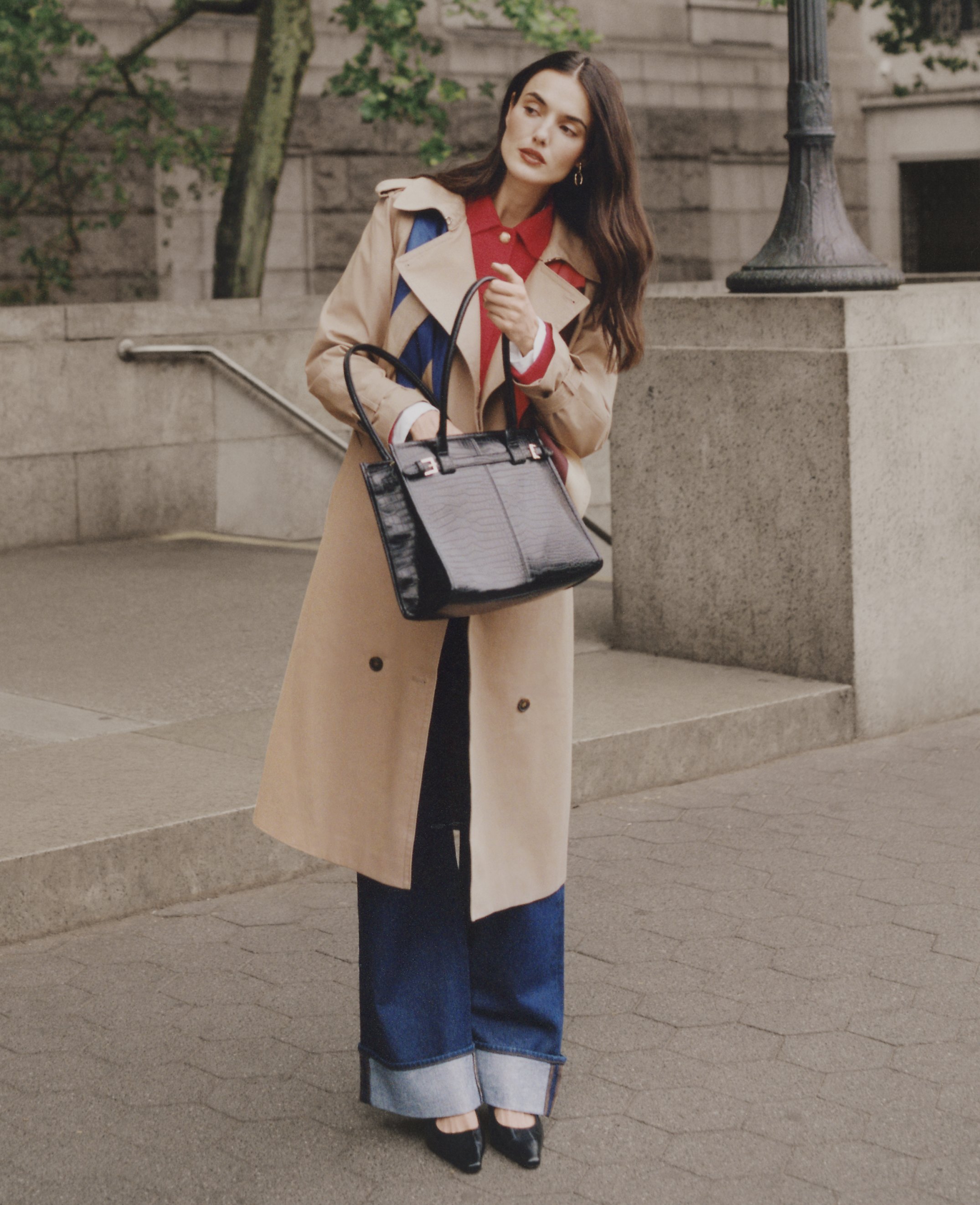 A woman in a trench coat and wide-leg jeans stands with a handbag in front of a stone staircase and lamppost.