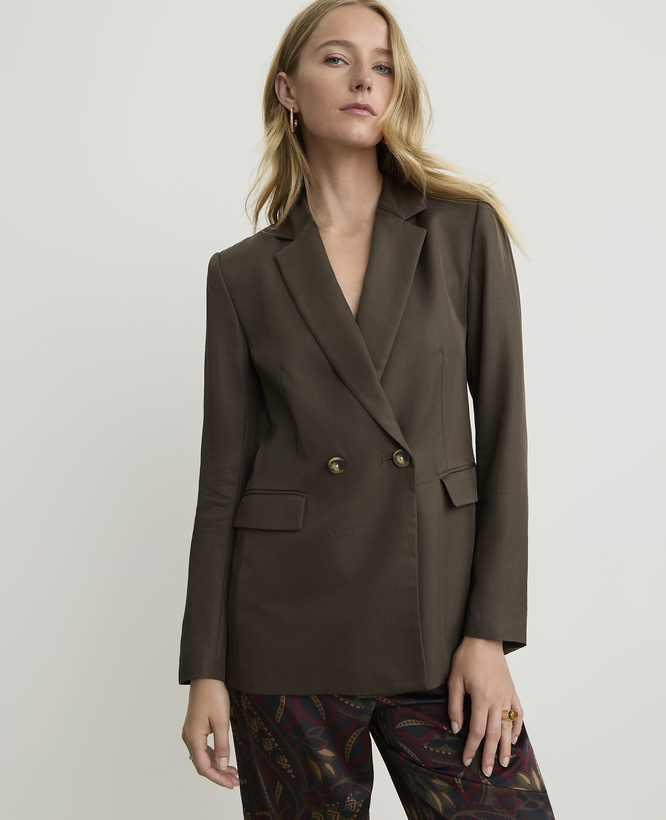 A woman in a dark brown double-breasted blazer and patterned pants stands against a plain white background.