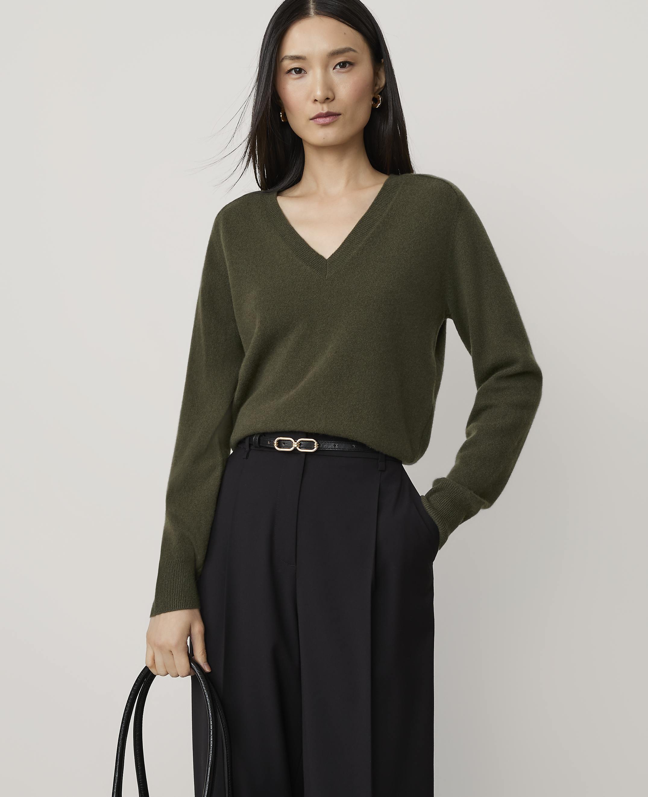 A woman in a green v-neck sweater and black pants stands against a plain, light background.