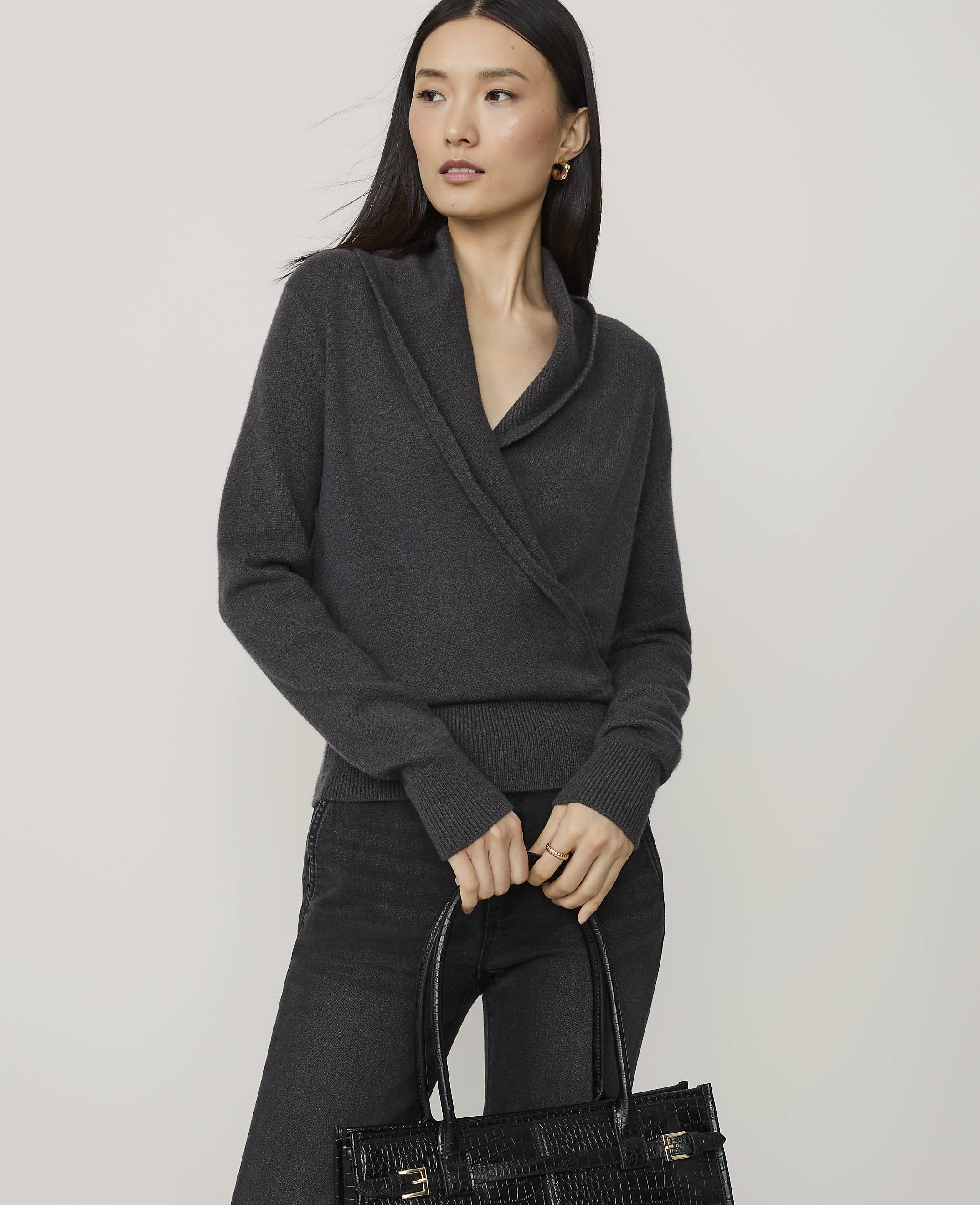 A woman in a dark grey wrap sweater and black jeans holds a black textured tote bag in front of a plain grey background.