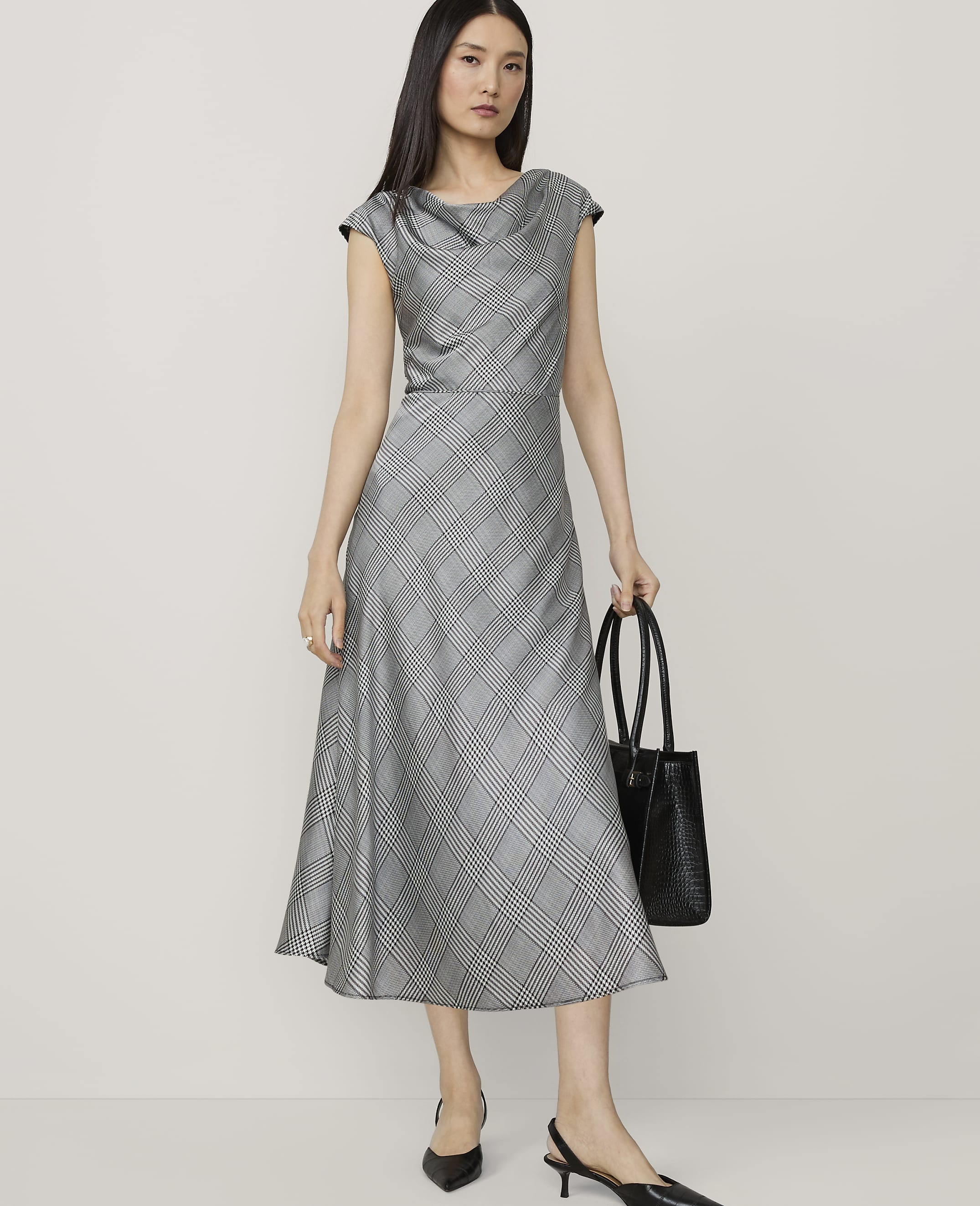 A woman in a grey plaid dress and black heels stands with a black tote bag against a plain, light grey background.