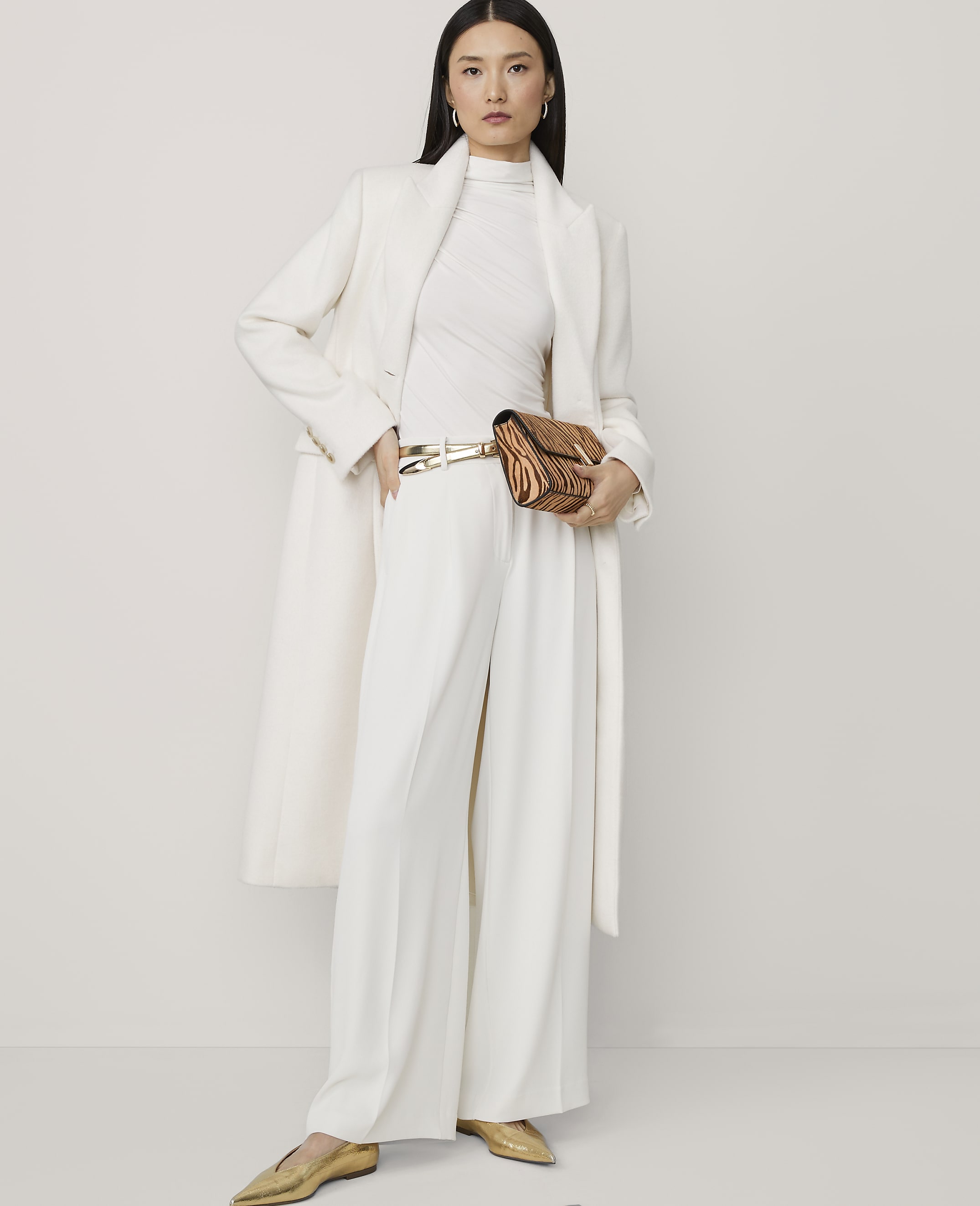 A woman in a white coat and pants poses against a plain white background, holding a patterned clutch.
