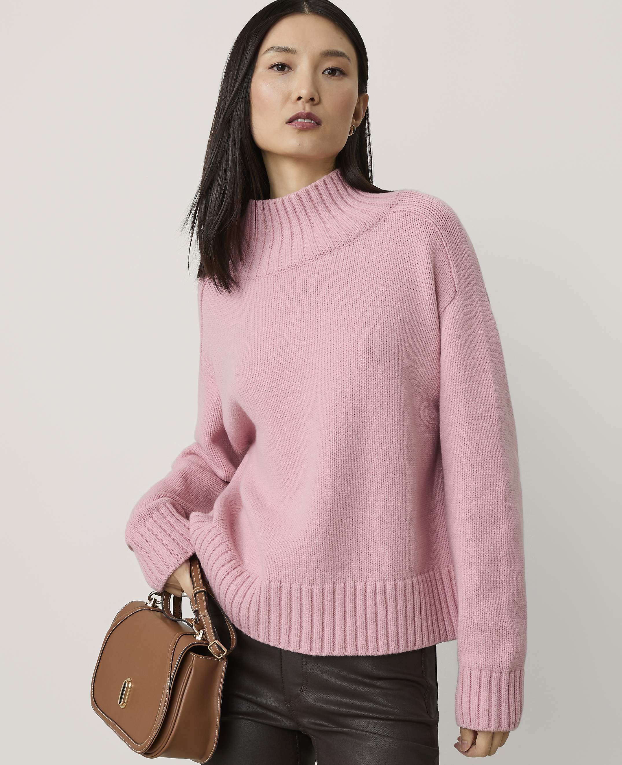 A woman in a pink sweater and dark pants holds a brown purse in front of a plain background.