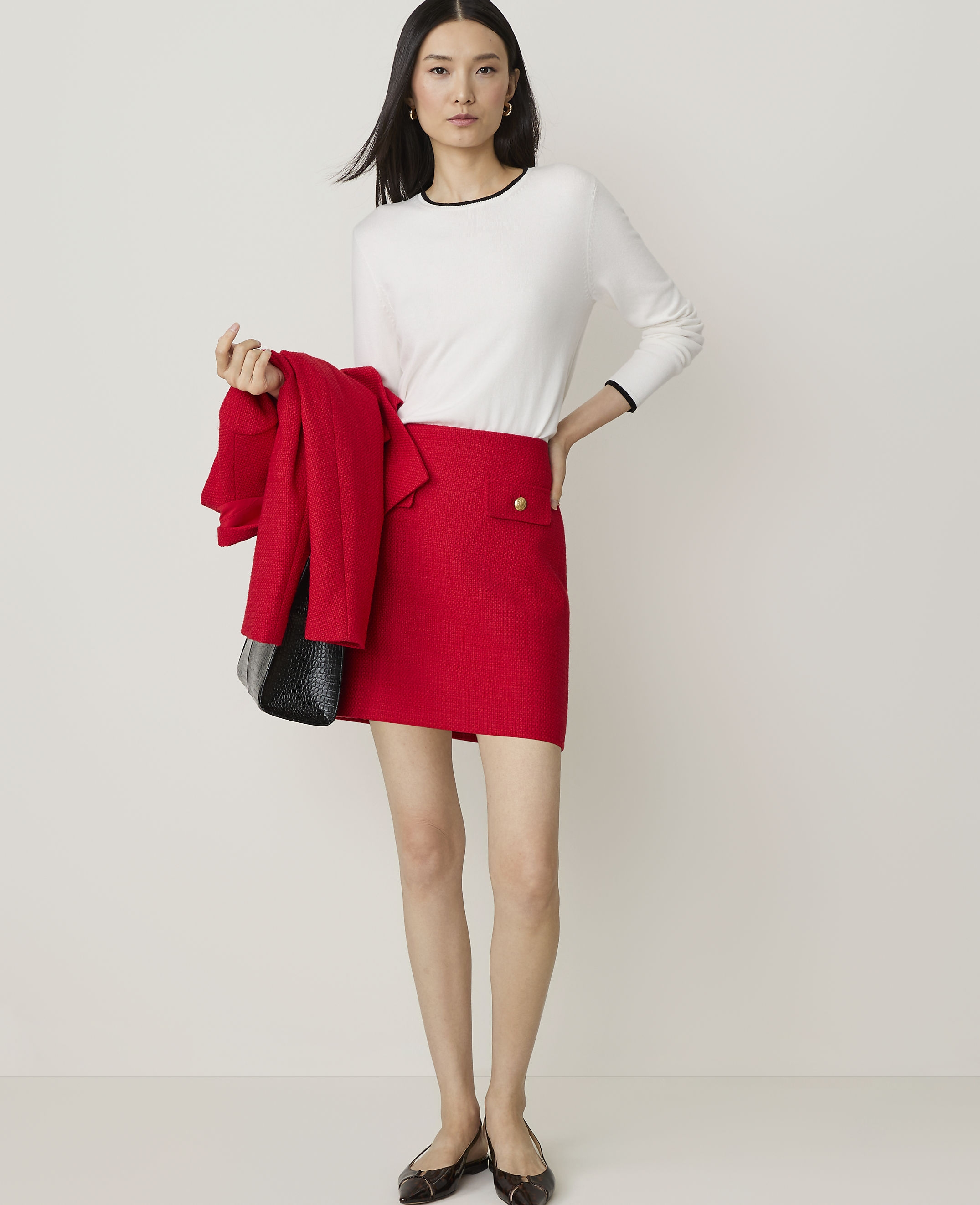 A woman in a red tweed skirt suit and black heels stands holding a black handbag against a plain white background.