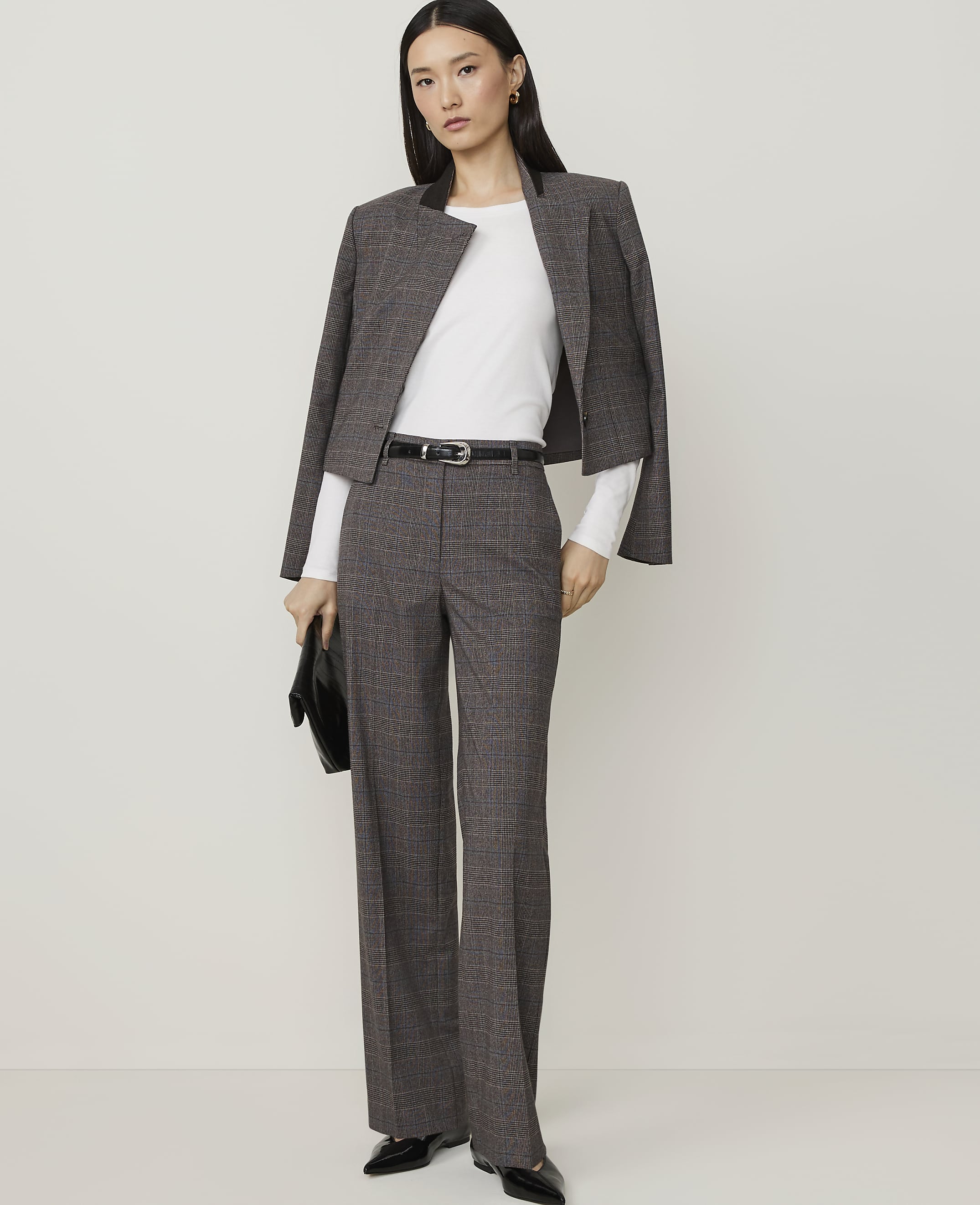 A woman in a grey plaid suit and black heels stands holding a dark woven clutch against a plain white background.