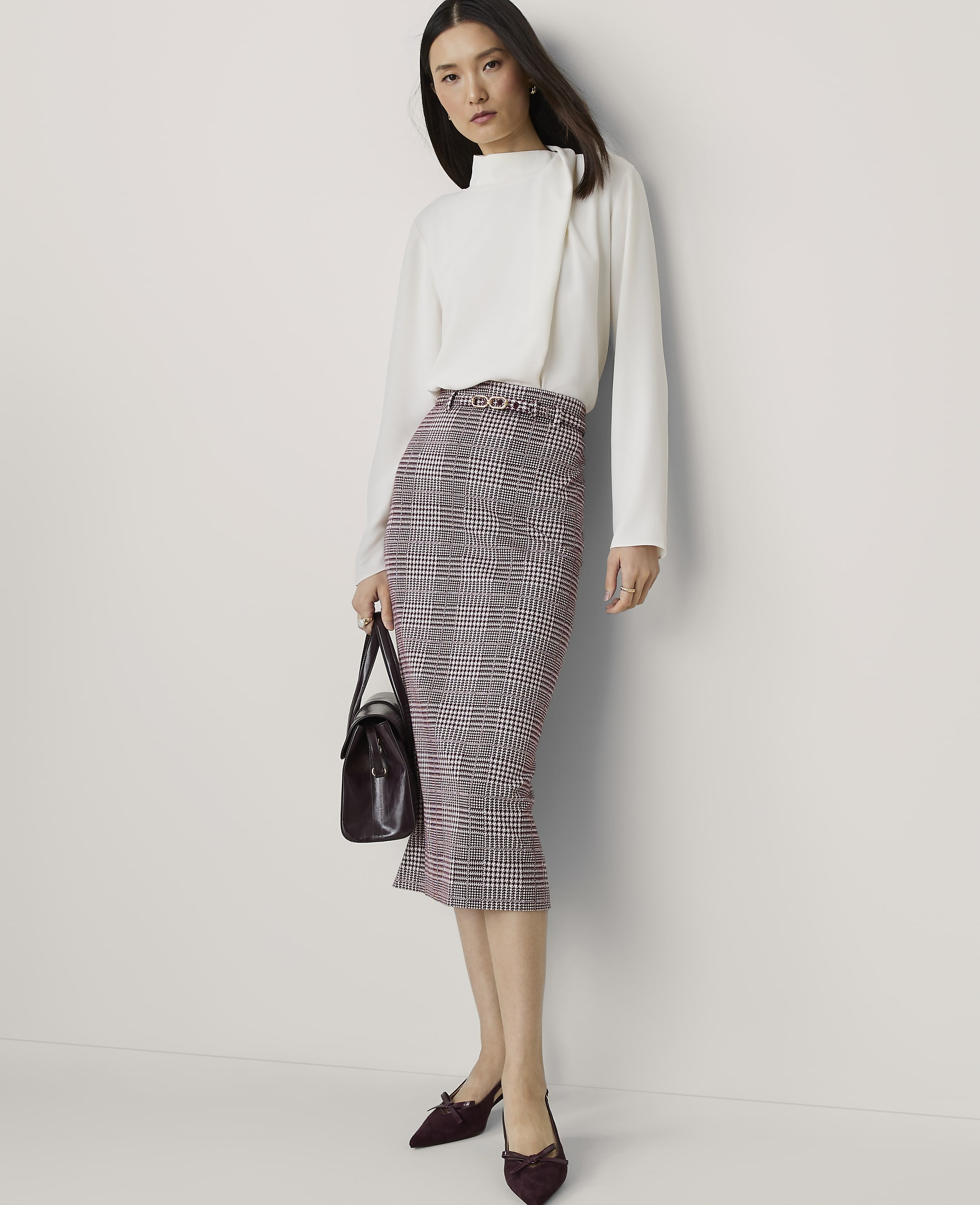 A woman in a white blouse and plaid skirt stands against a plain white background, holding a dark handbag.