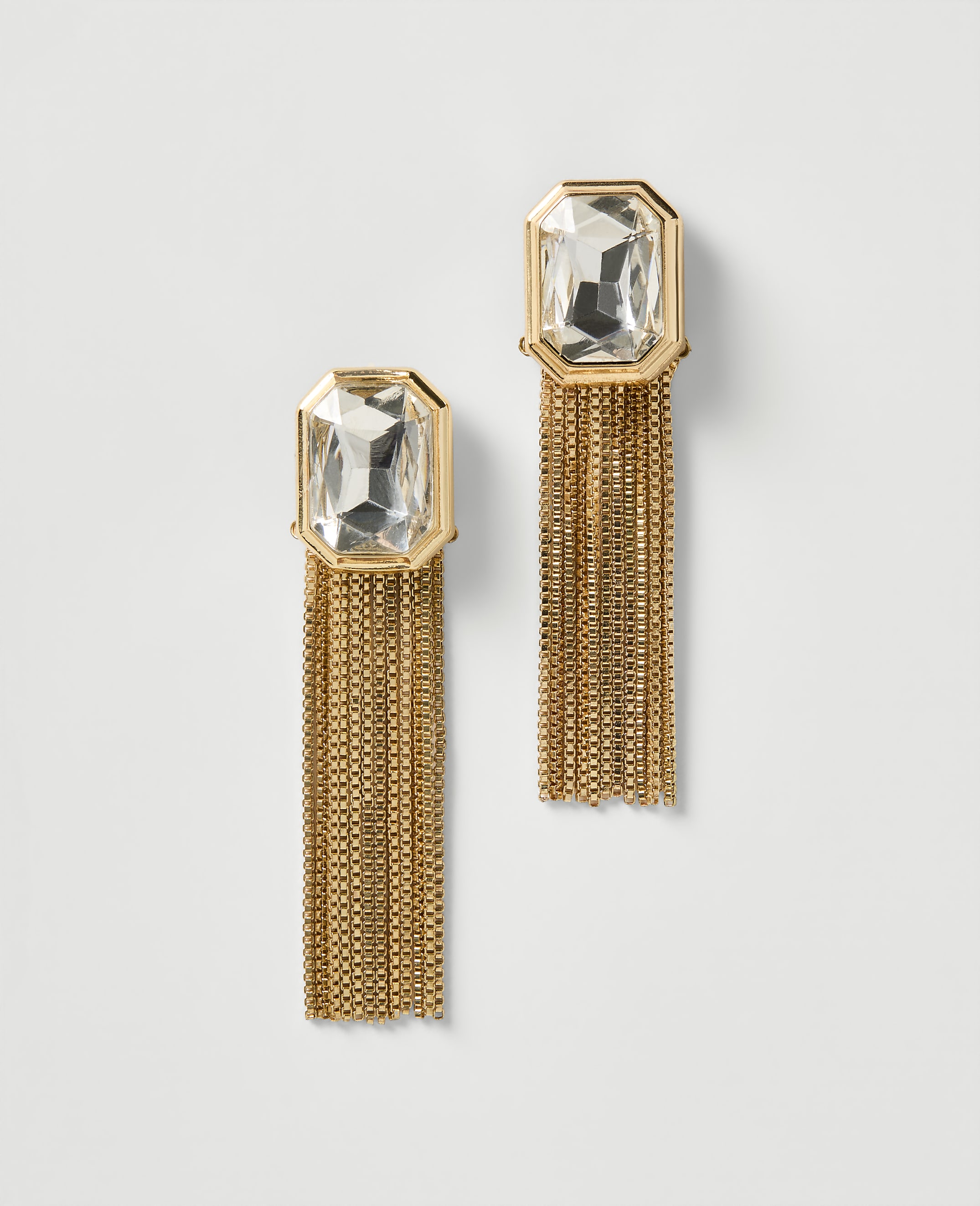 A pair of gold earrings with clear rectangular gems and long, shimmering gold chains hang against a plain white background.