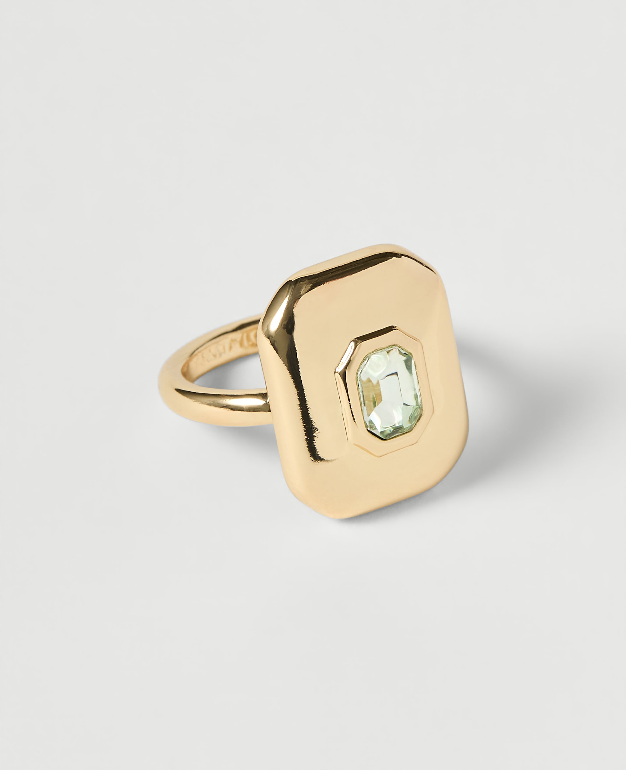 A gold ring with a rectangular face and a light green gemstone sits on a plain white background.