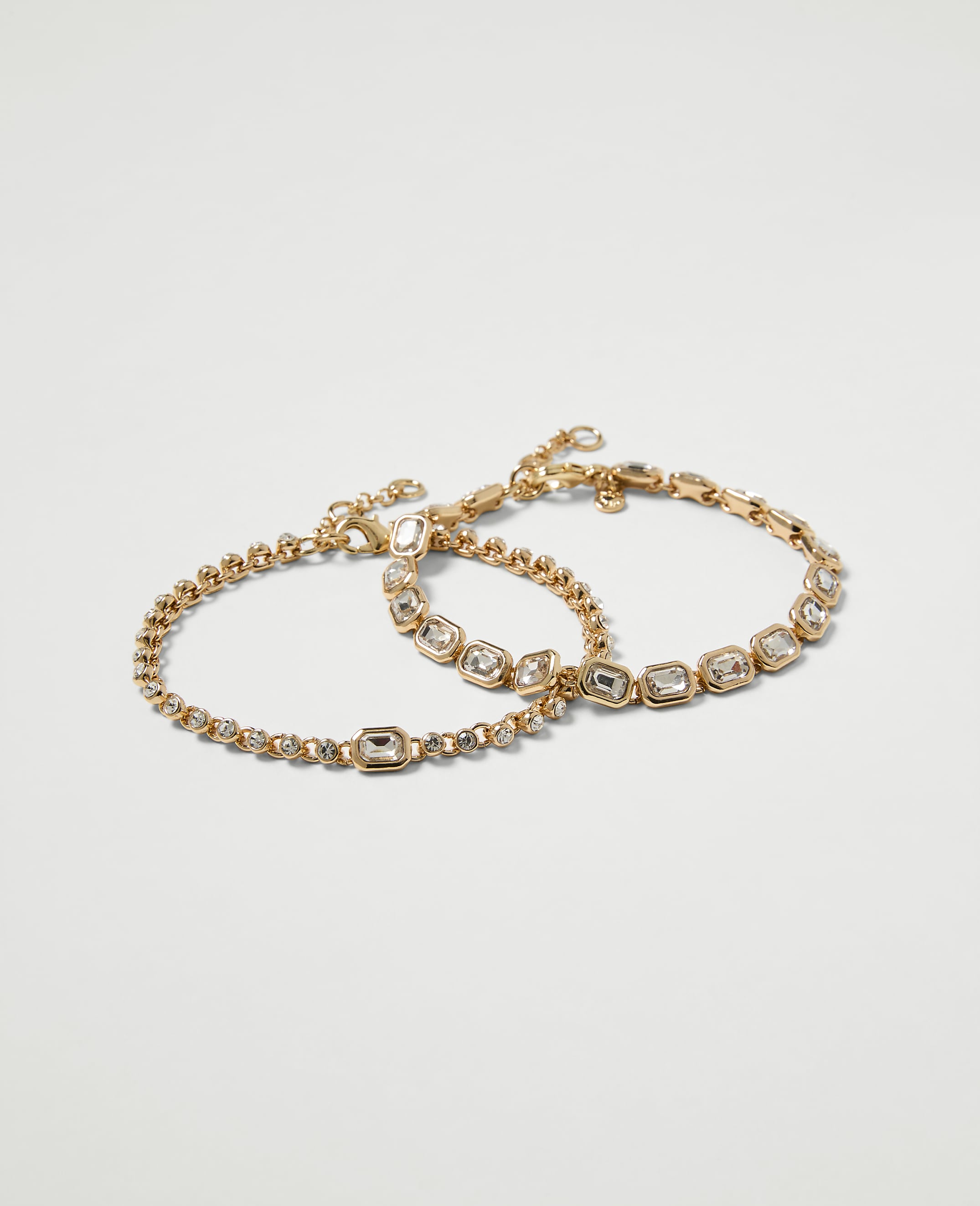 Two delicate gold bracelets, one with a simple chain and the other adorned with clear stones, rest against a plain white background.