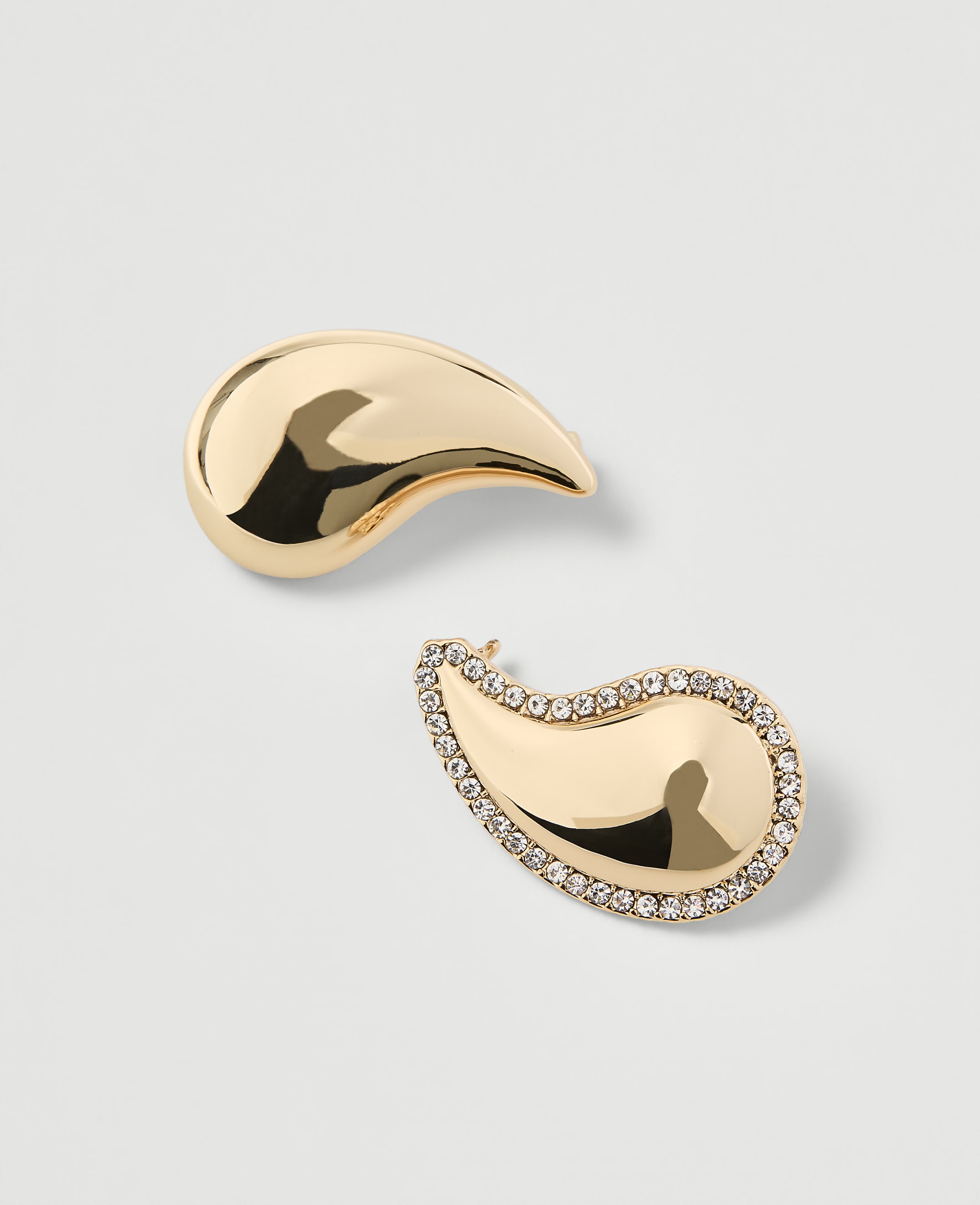 Two gold paisley-shaped earrings, one plain and the other encrusted with small diamonds, rest on a plain white background.