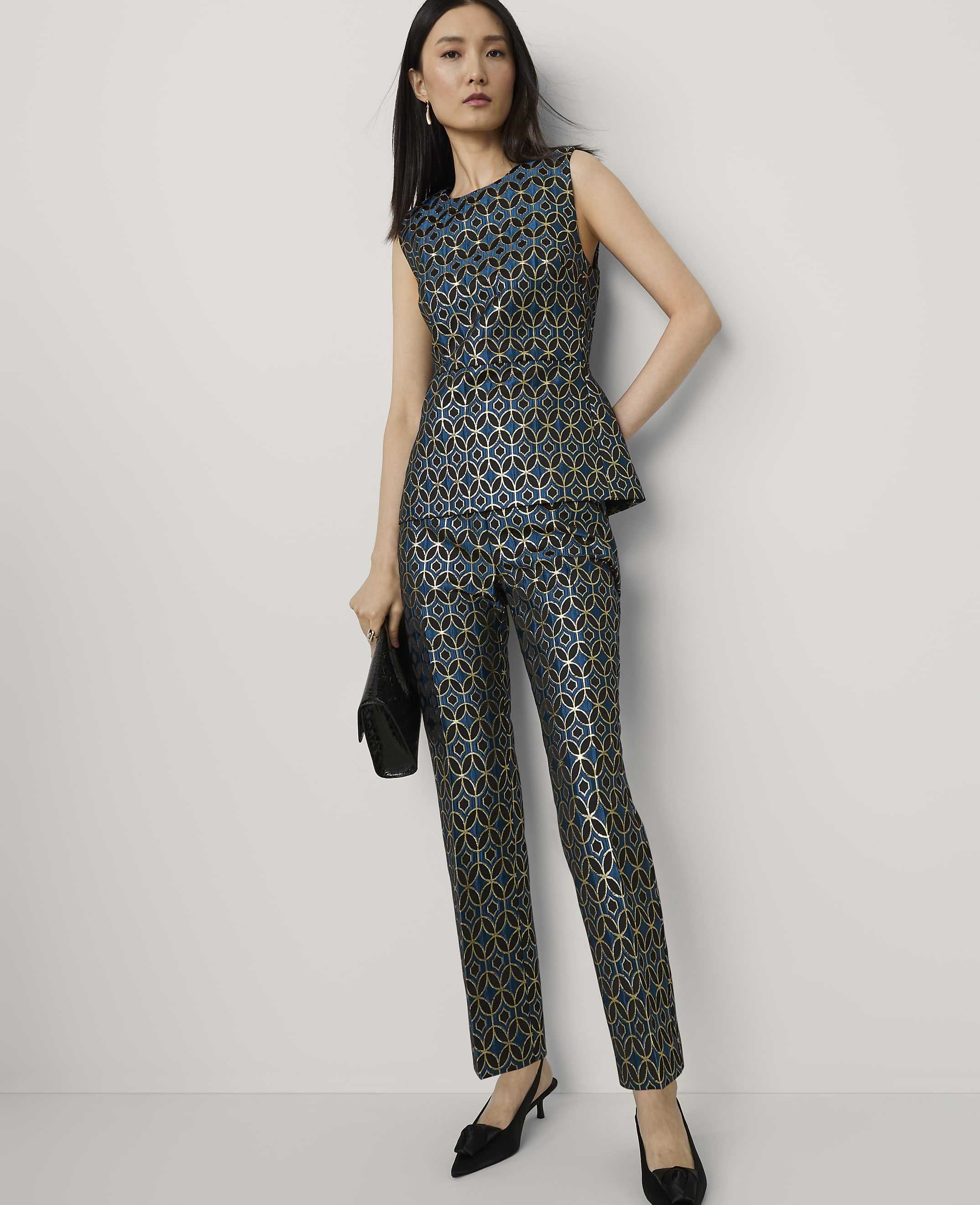 A woman in a teal and gold patterned peplum top and matching pants stands with a black clutch against a plain white background.