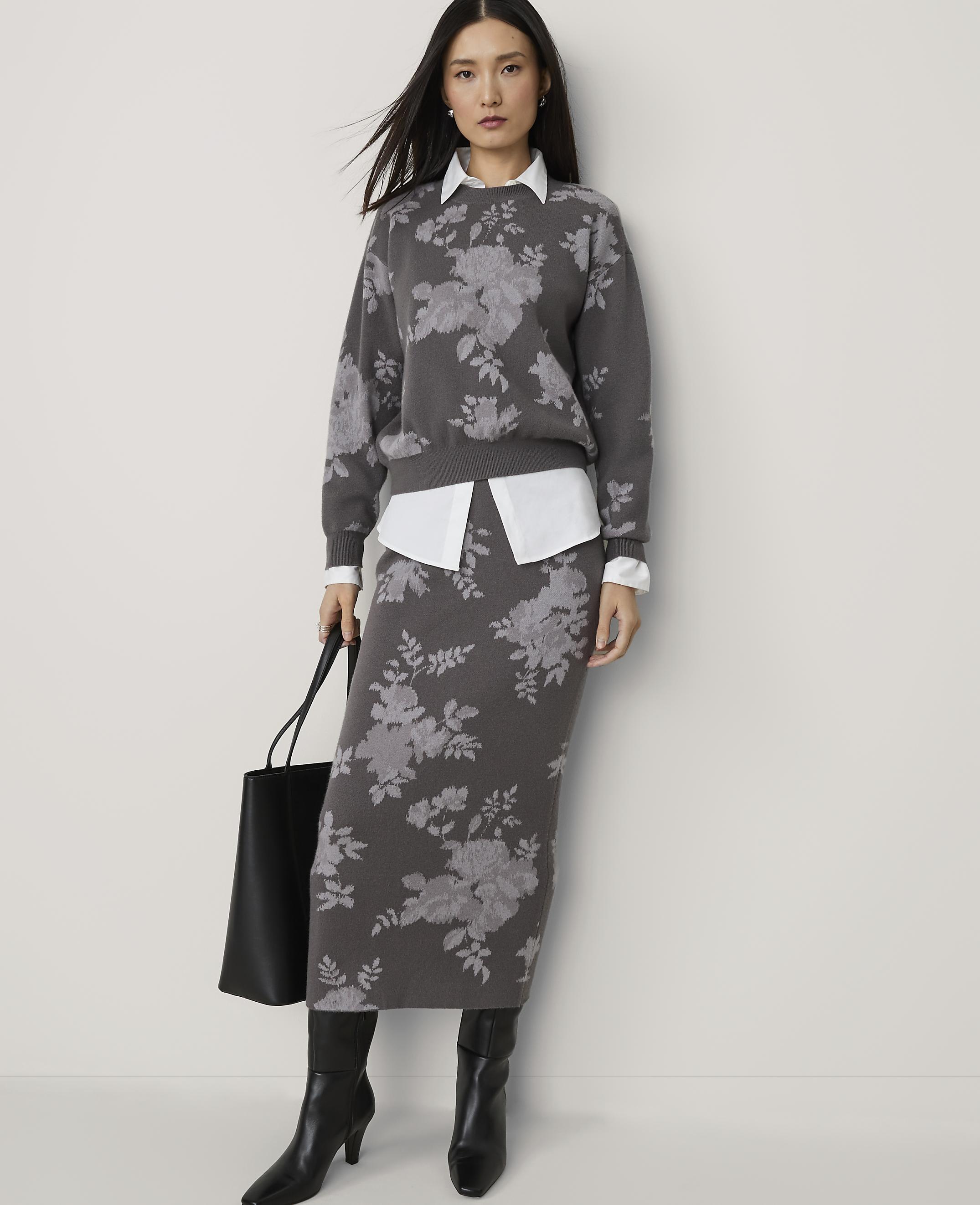 A woman in a floral sweater and skirt stands against a plain background, holding a black tote bag.