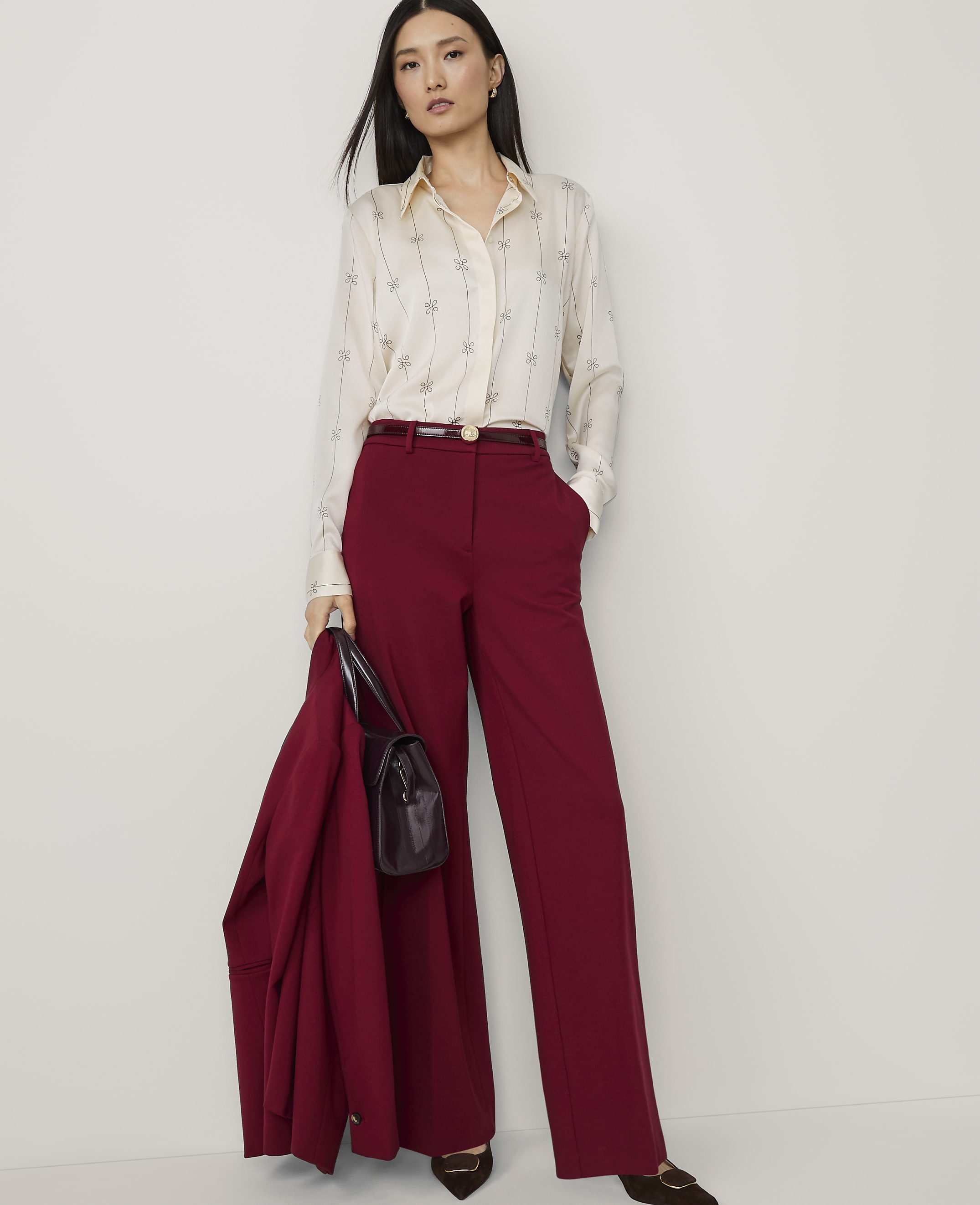 A woman in a cream patterned blouse and maroon wide-leg pants stands against a plain white background, holding a maroon jacket and handbag.